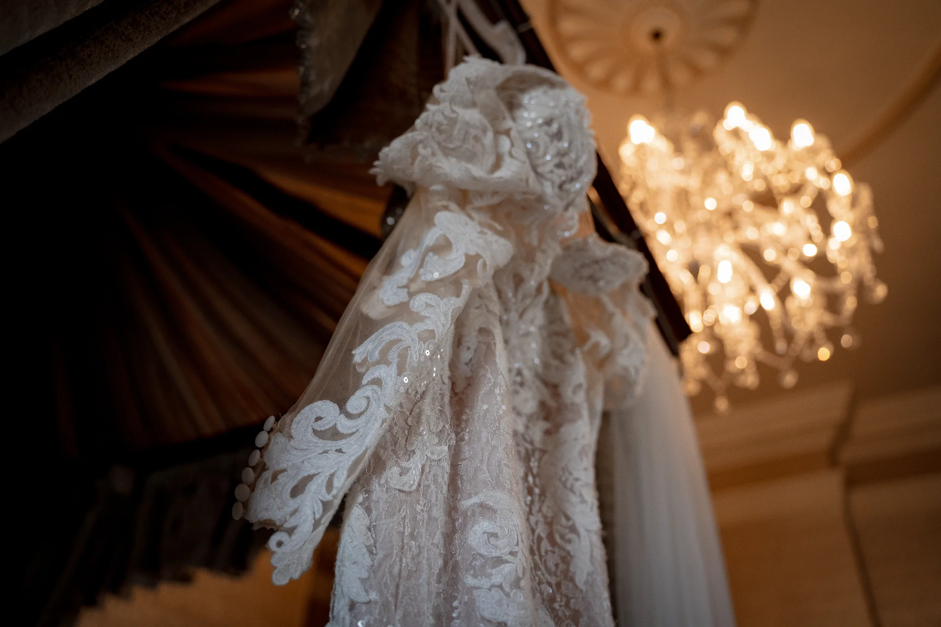 An intricately detailed lace wedding dress hangs in an elegant room, illuminated by a warm, glowing chandelier in the background.