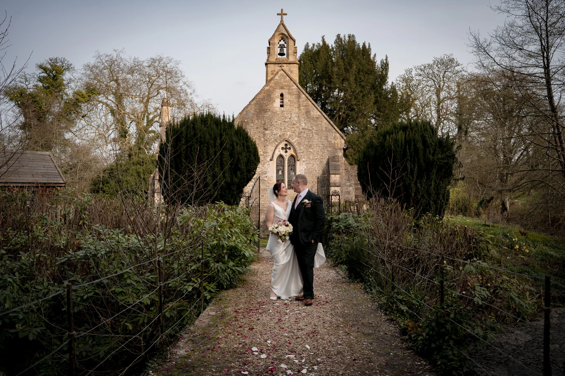 A bride and groom walk hand in hand down a petal-strewn path towards an historic stone church on their wedding day.