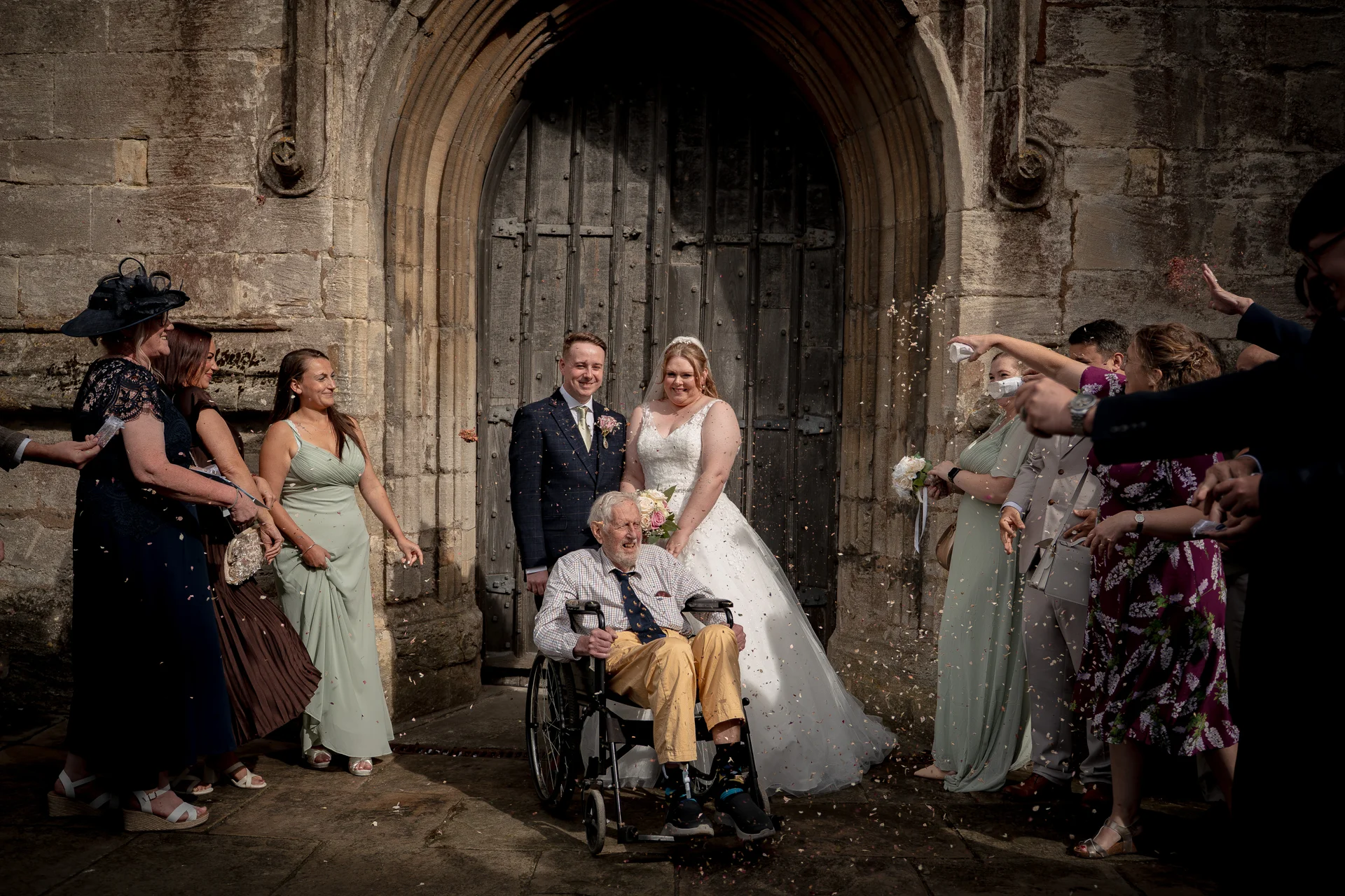 Newlyweds stand outside a historic stone church with large wooden doors as guests shower them and an elderly guest in a wheelchair with confetti.