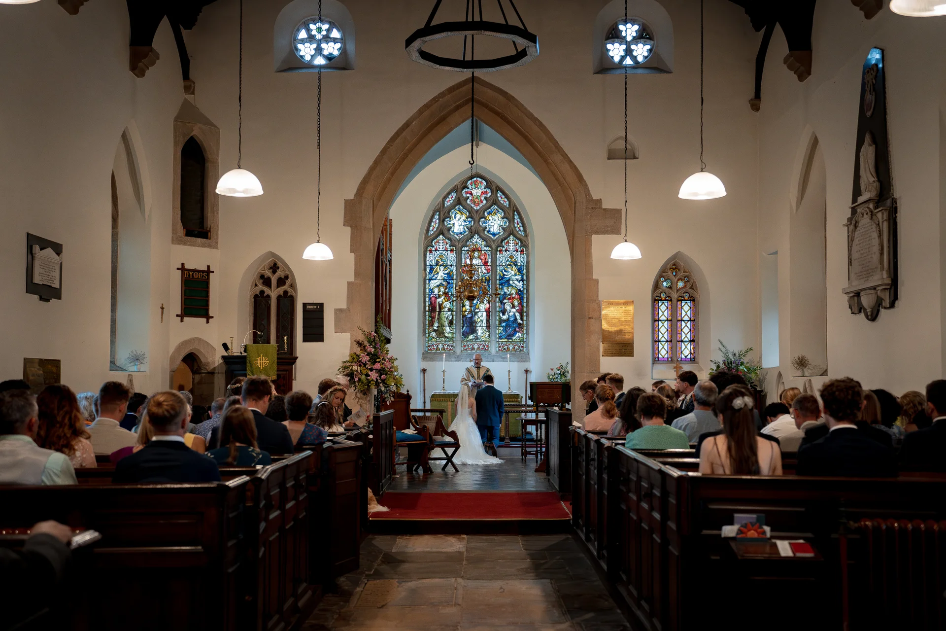 A wide-angle documentary shot of a church wedding ceremony, showing the bride and groom kneeling at the altar framed by a grand stone archway and colourful stained glass windows.
