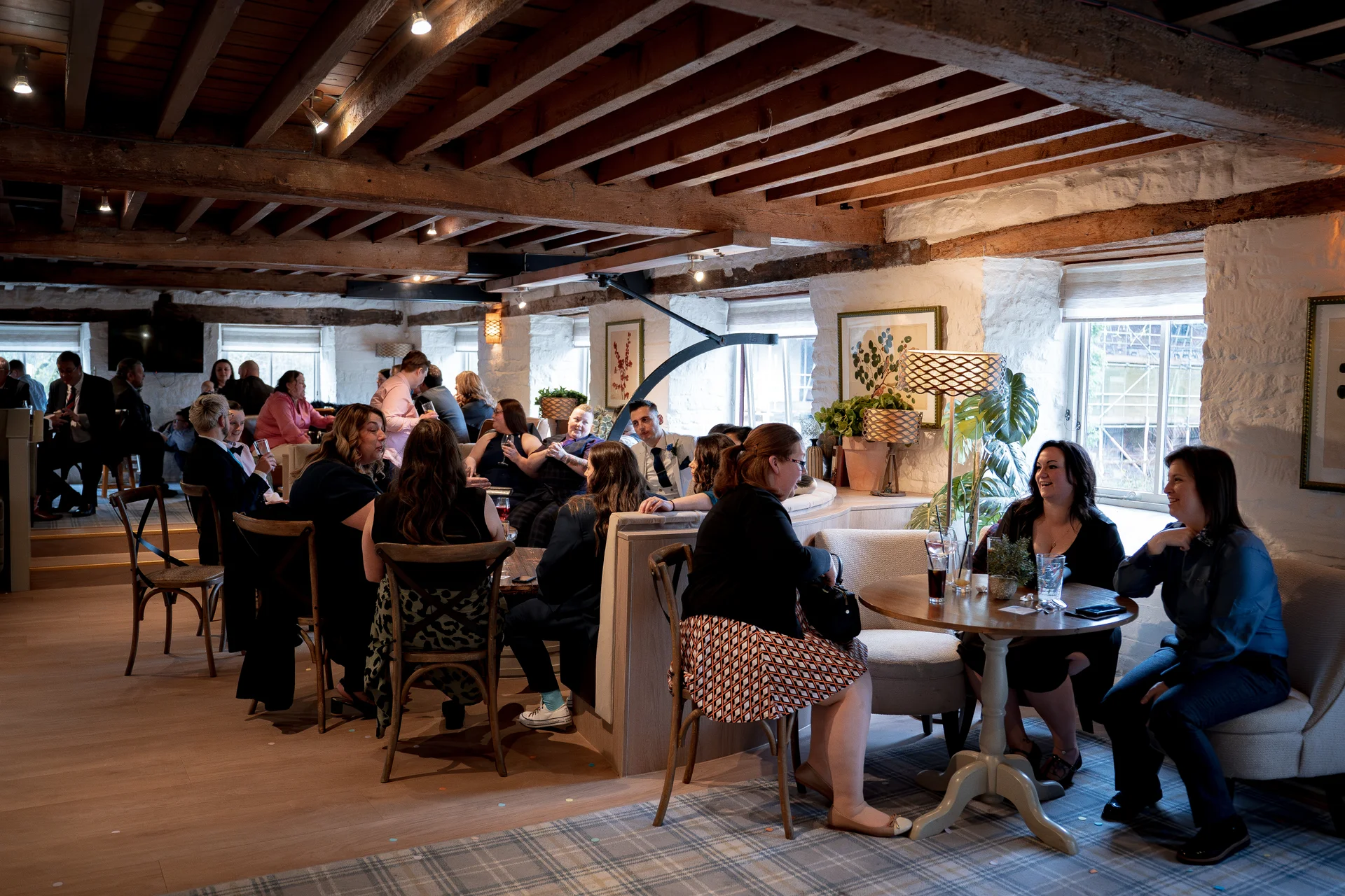 Wedding guests mingle and converse in a rustic barn venue featuring exposed wooden ceiling beams and whitewashed stone walls.