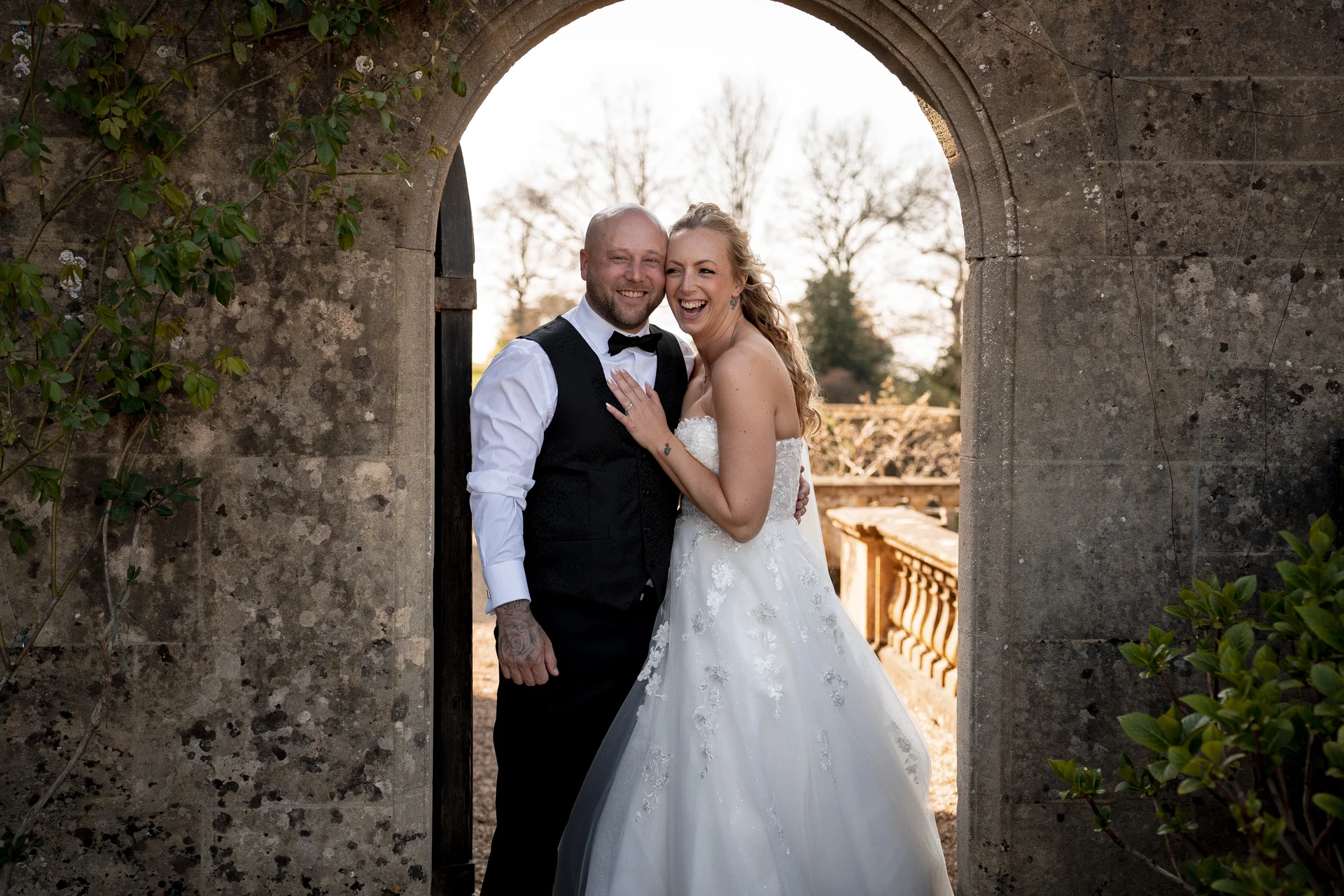 A newlywed couple laughing joyfully together while posing inside a rustic stone archway framed by climbing foliage.