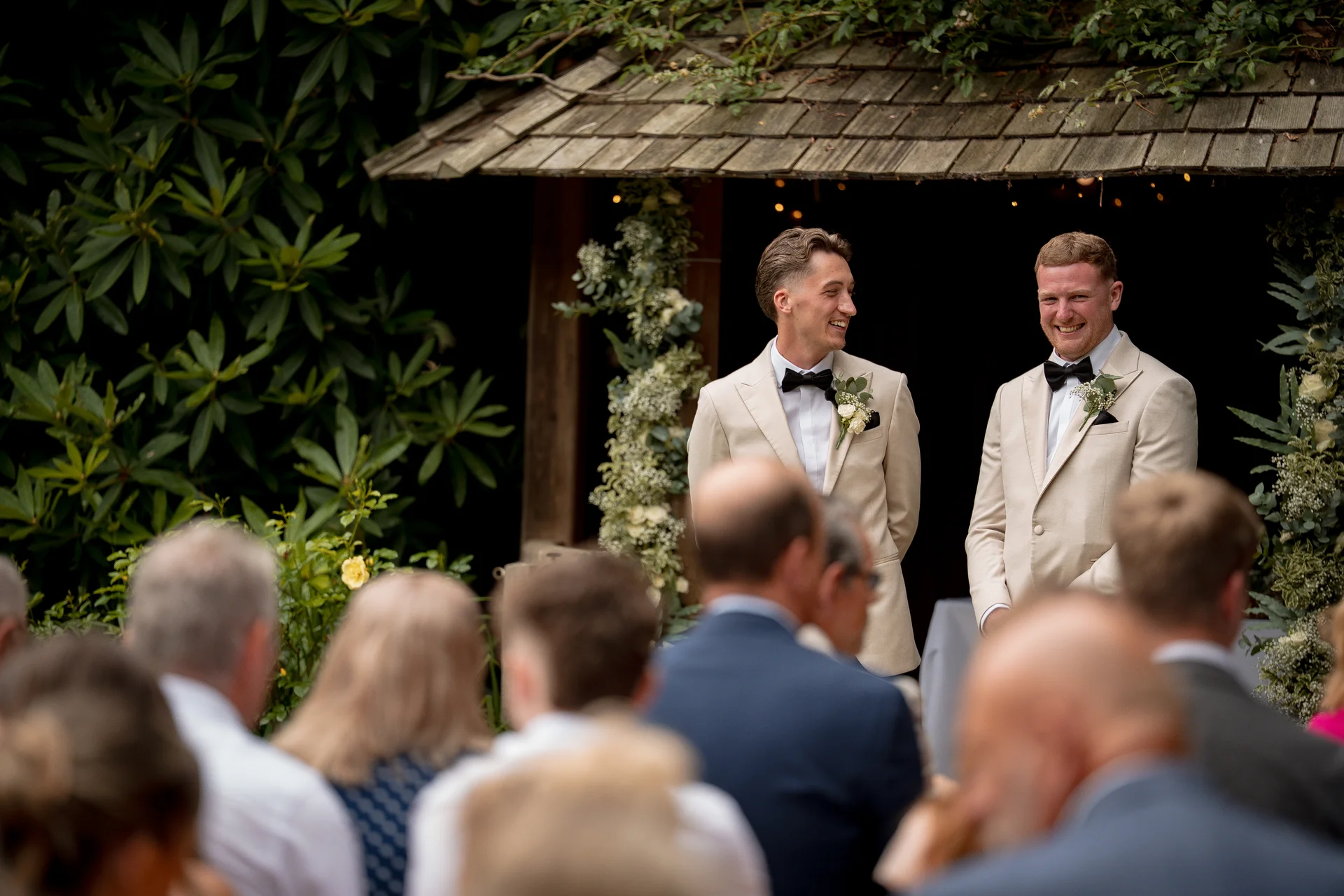 Two smiling grooms in matching beige suits and black bow ties stand at the altar during an outdoor woodland wedding ceremony, viewed past blurred seated guests.