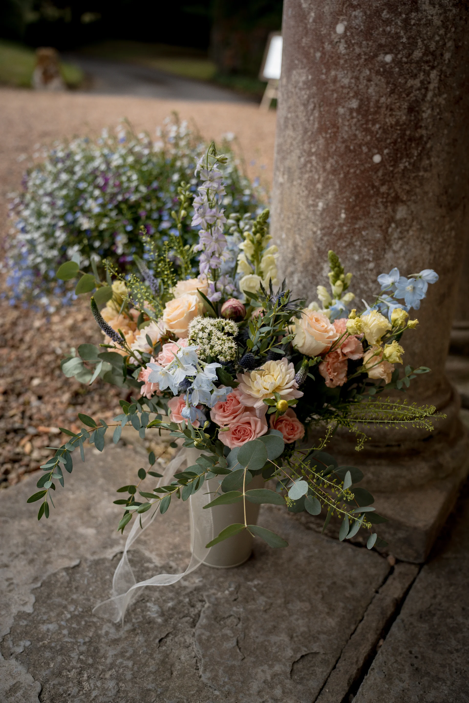 A lush hand-tied bridal bouquet featuring soft peach roses, blue delphiniums, and trailing eucalyptus rests against a weathered stone pillar.