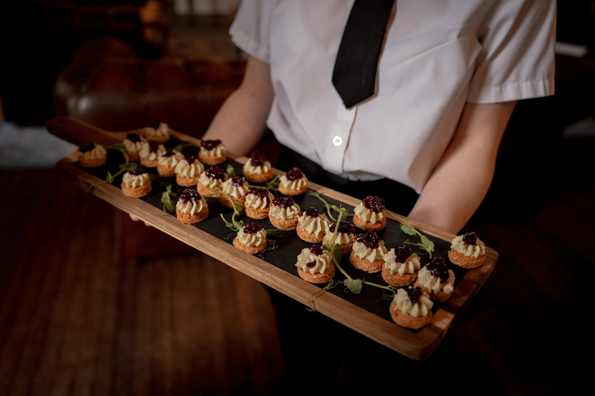 A member of the waitstaff in a crisp white shirt and black tie presents a wooden board laden with gourmet canapes featuring piped toppings and dark fruit compote at a wedding reception.