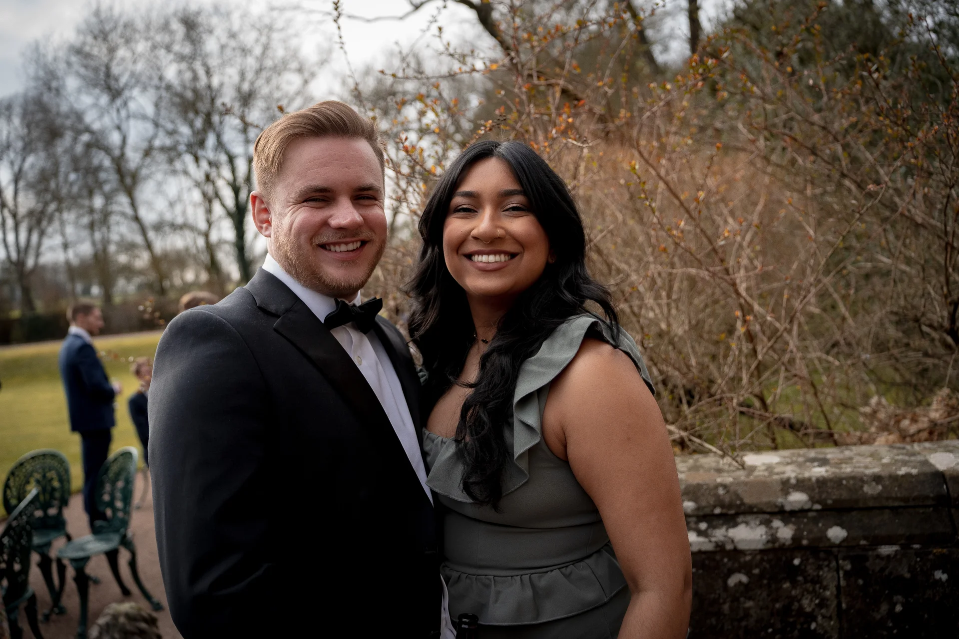 An interracial couple smiling happily for an outdoor portrait at a wedding, with the groom wearing a black tuxedo and the guest wearing a sage green dress.
