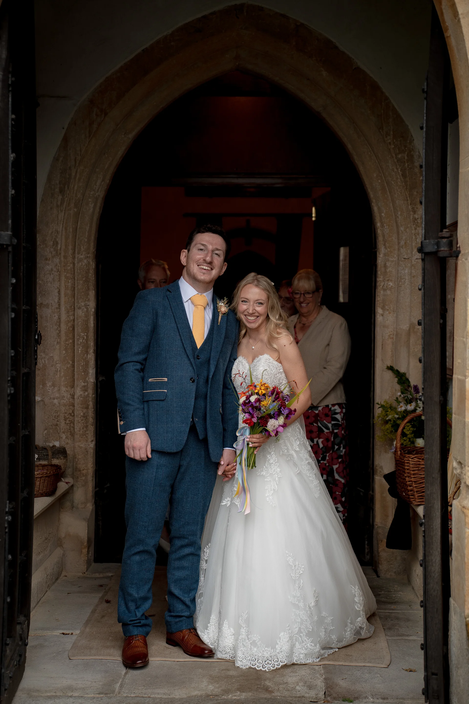 A newlywed couple smiles broadly while holding hands as they exit a historic stone church doorway.
