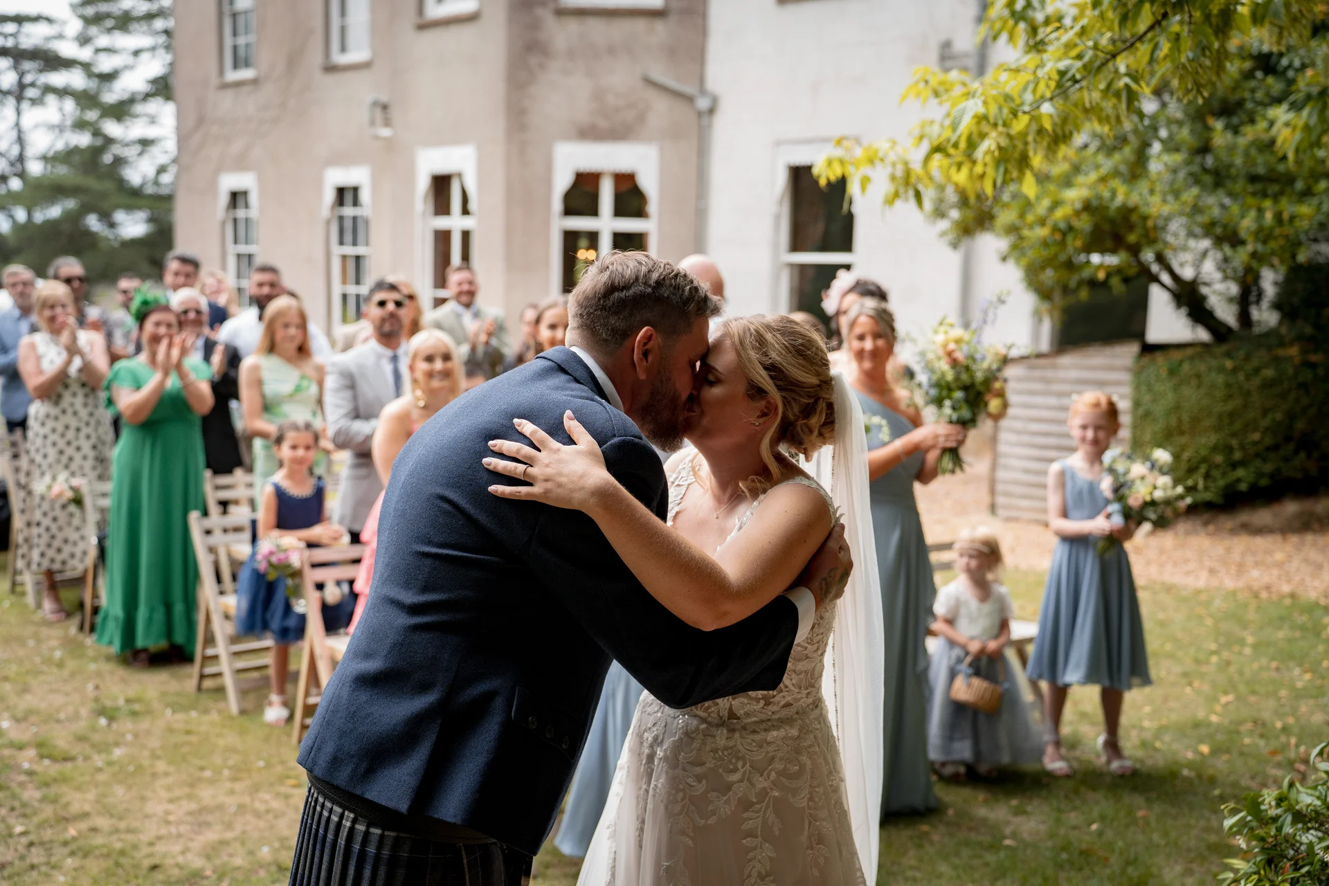 Newlywed bride and groom share their first kiss during an outdoor garden ceremony while guests applaud in the background.