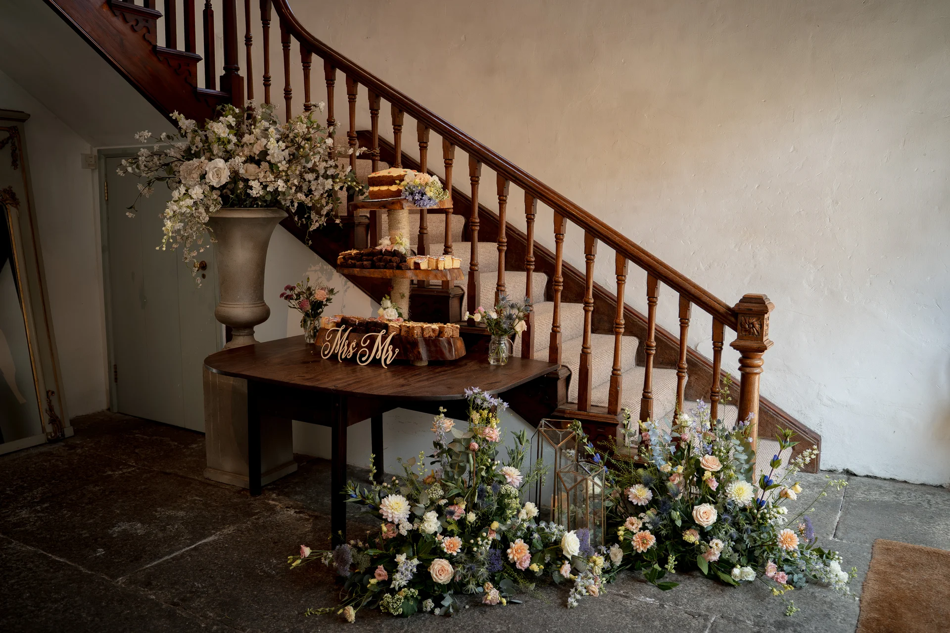 A rustic brownie stack wedding cake sits on a wooden table adorned with a Mrs & Mr sign, placed in front of a grand wooden staircase decorated with lush floral arrangements and lanterns.
