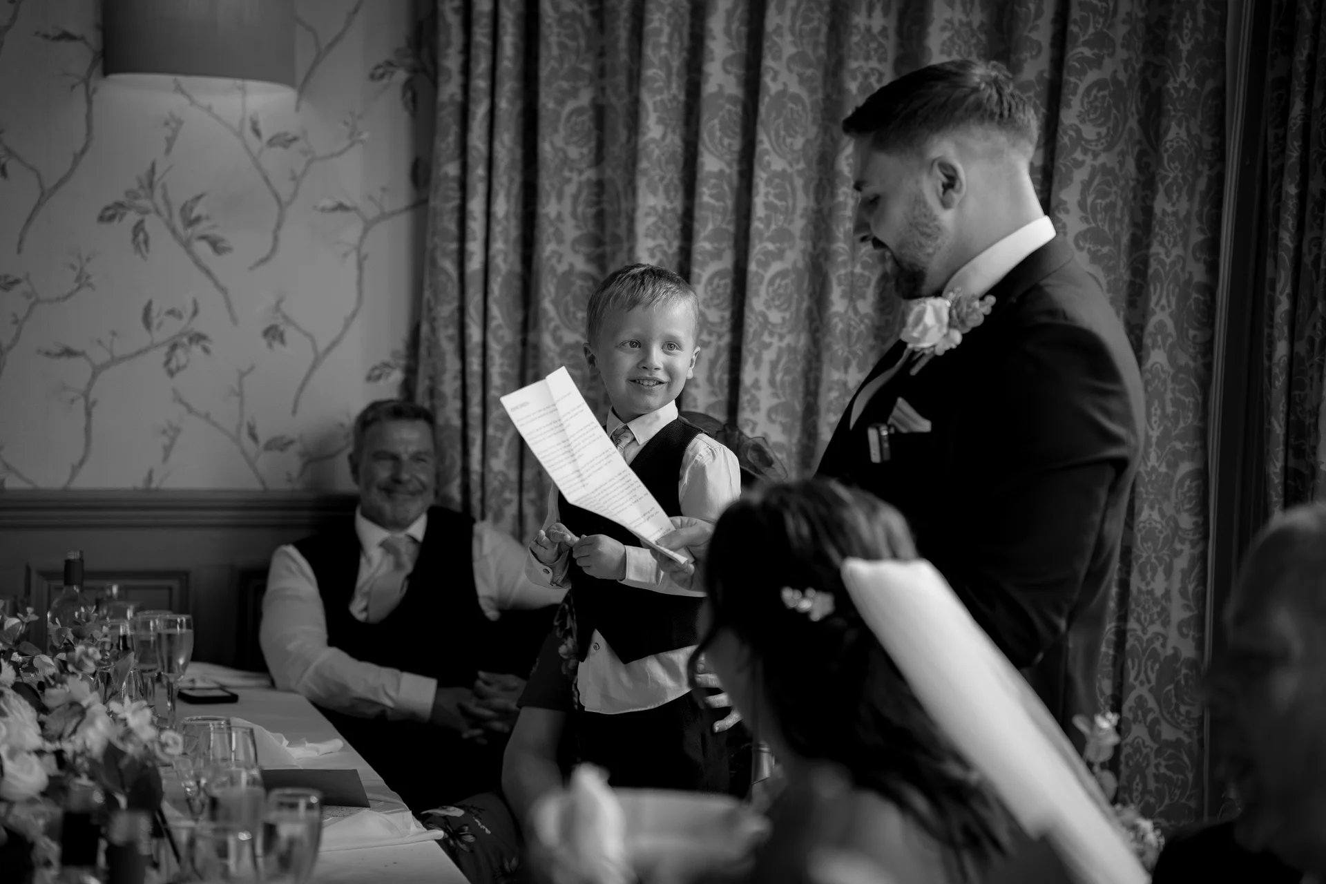 A young page boy happily holds up a speech while standing near the groom at the top table during the wedding reception.