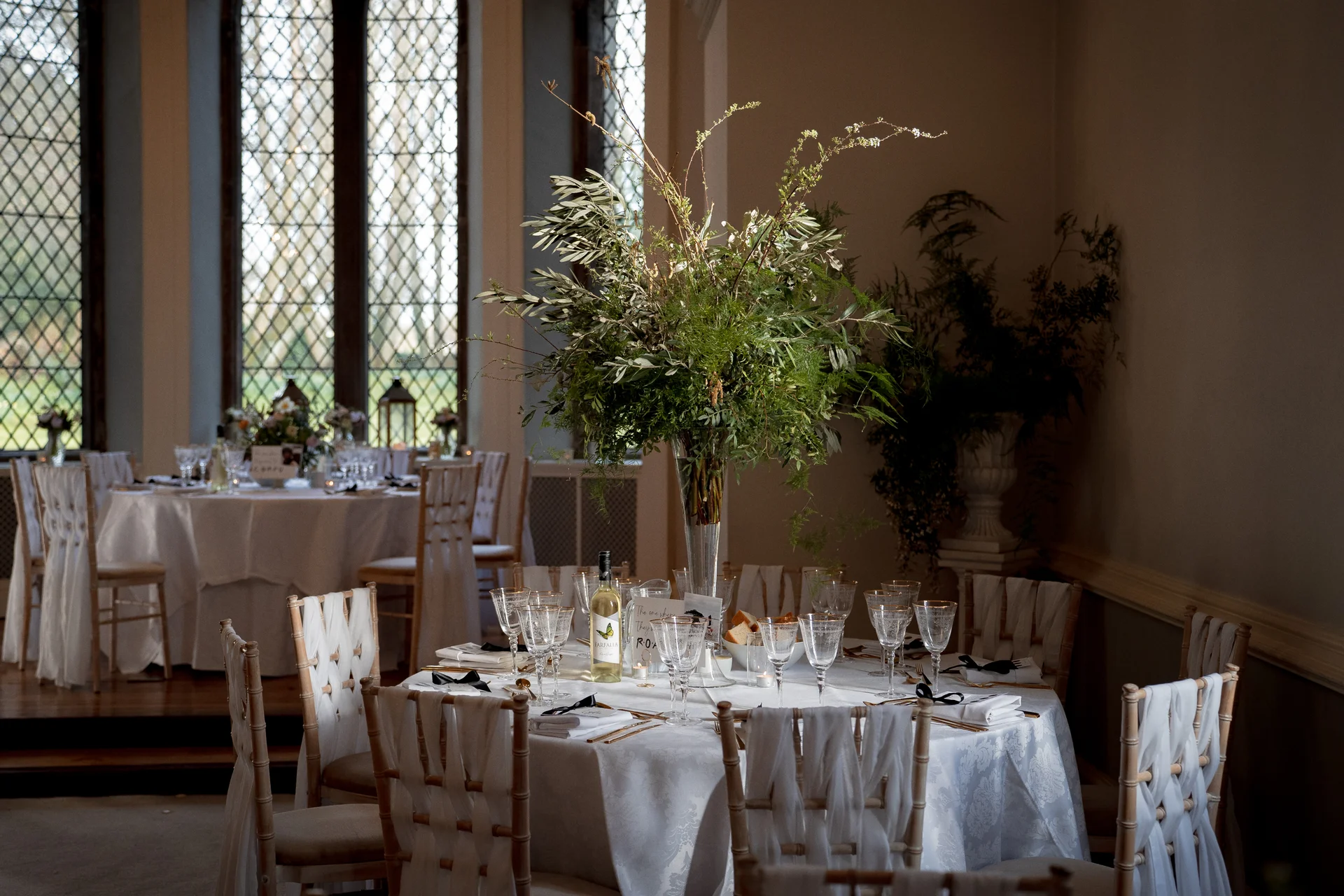 An elegant wedding reception table features a towering green foliage centrepiece in a glass vase, surrounded by white linens and crystal glassware in a light-filled historic room.