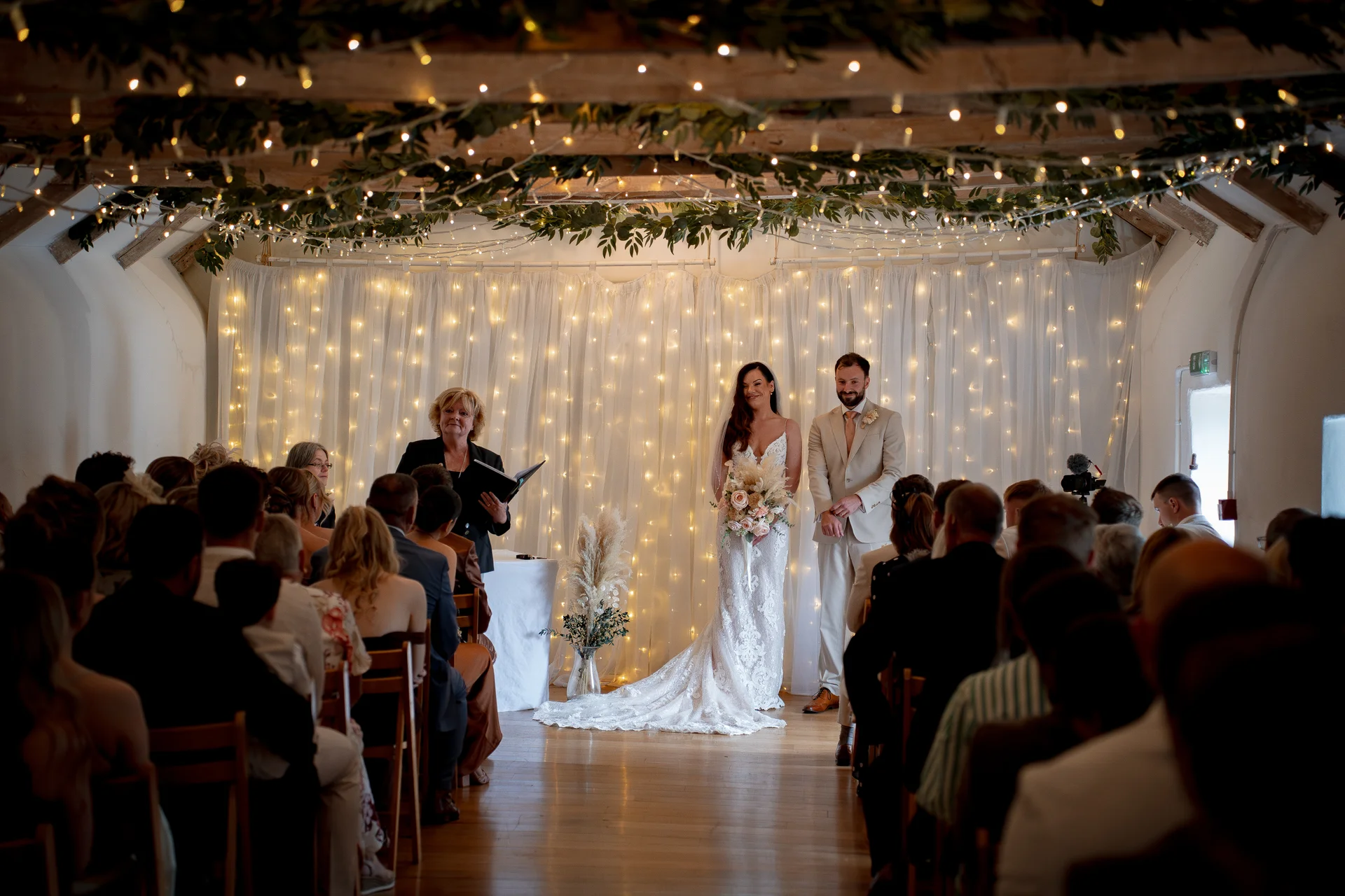 A bride holding a bouquet and a groom standing at the altar during an indoor barn ceremony illuminated by a glowing fairy light backdrop and ceiling canopy.