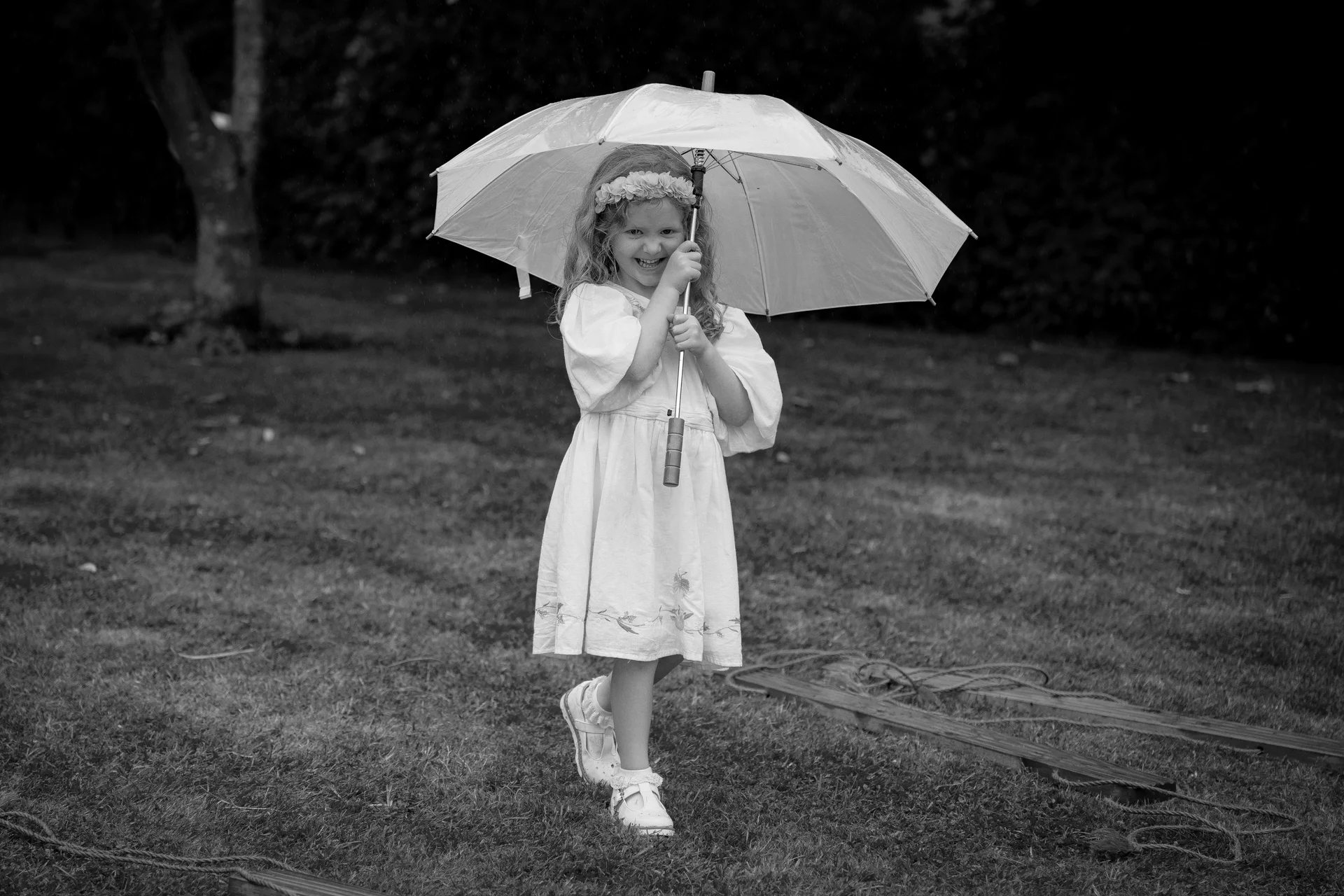 A young flower girl wearing a floral crown and white dress stands outdoors in the rain holding a clear umbrella.