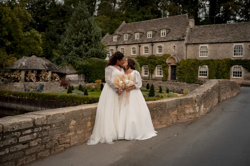 Two brides in white gowns share a tender kiss while holding dried flower bouquets on a stone bridge outside a historic country house venue.