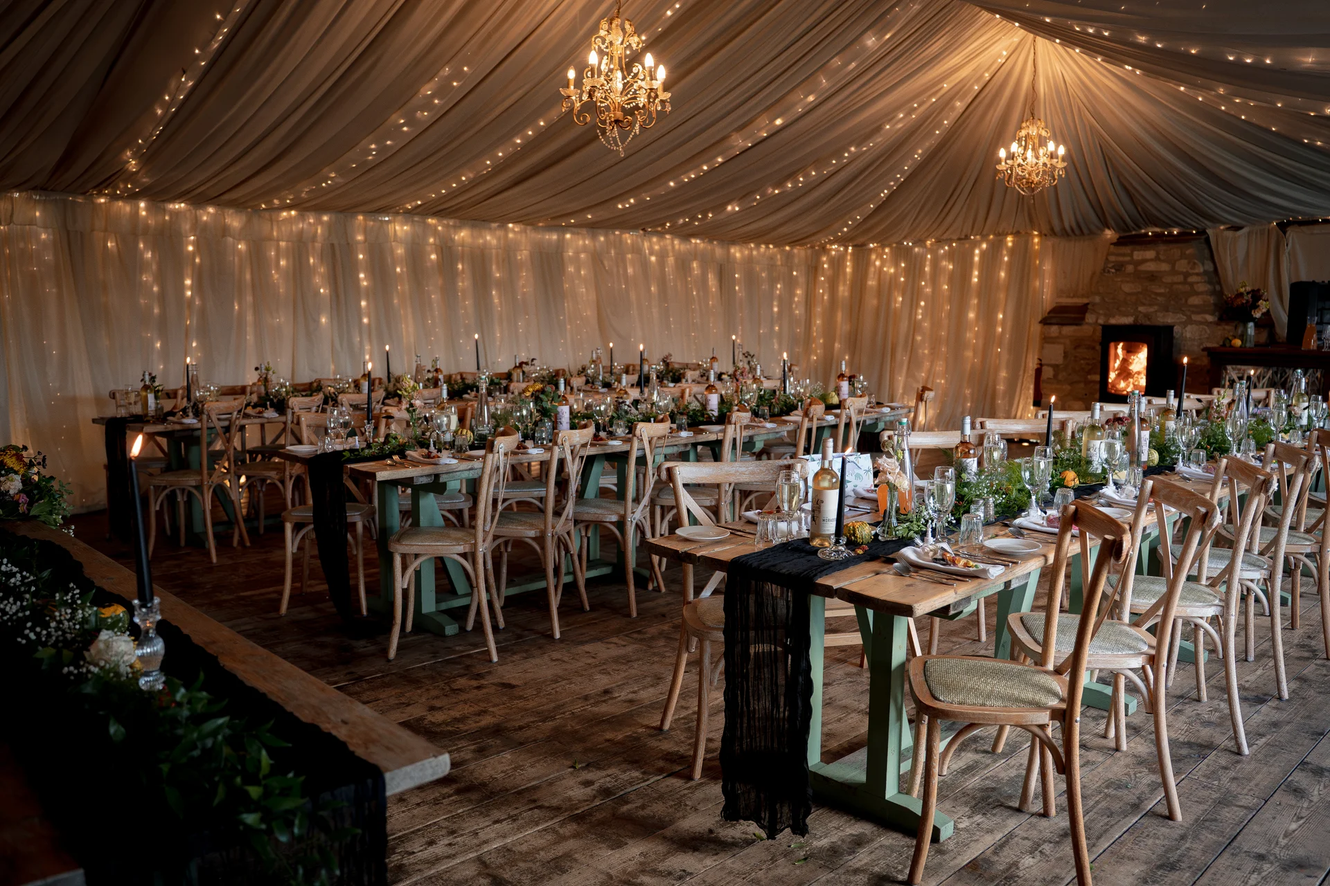 An elegantly decorated marquee wedding reception features long wooden banquet tables set with black runners, taper candles, and glittering fairy light canopies overhead.