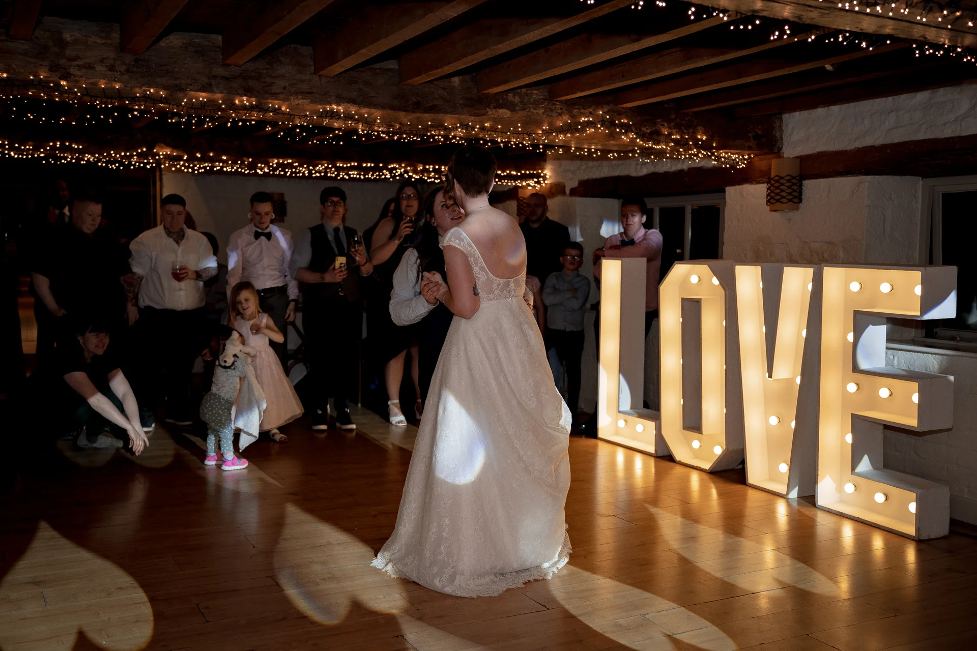 Two brides share their first dance on a wooden floor inside a rustic barn illuminated by fairy lights and giant illuminated love letters.