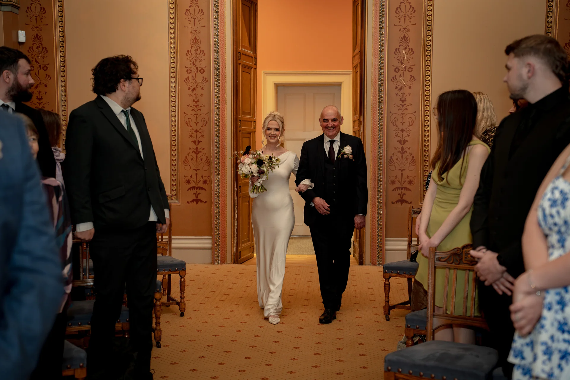 A smiling bride holding a bouquet walks down the aisle arm in arm with her father inside a grand, classically decorated venue.