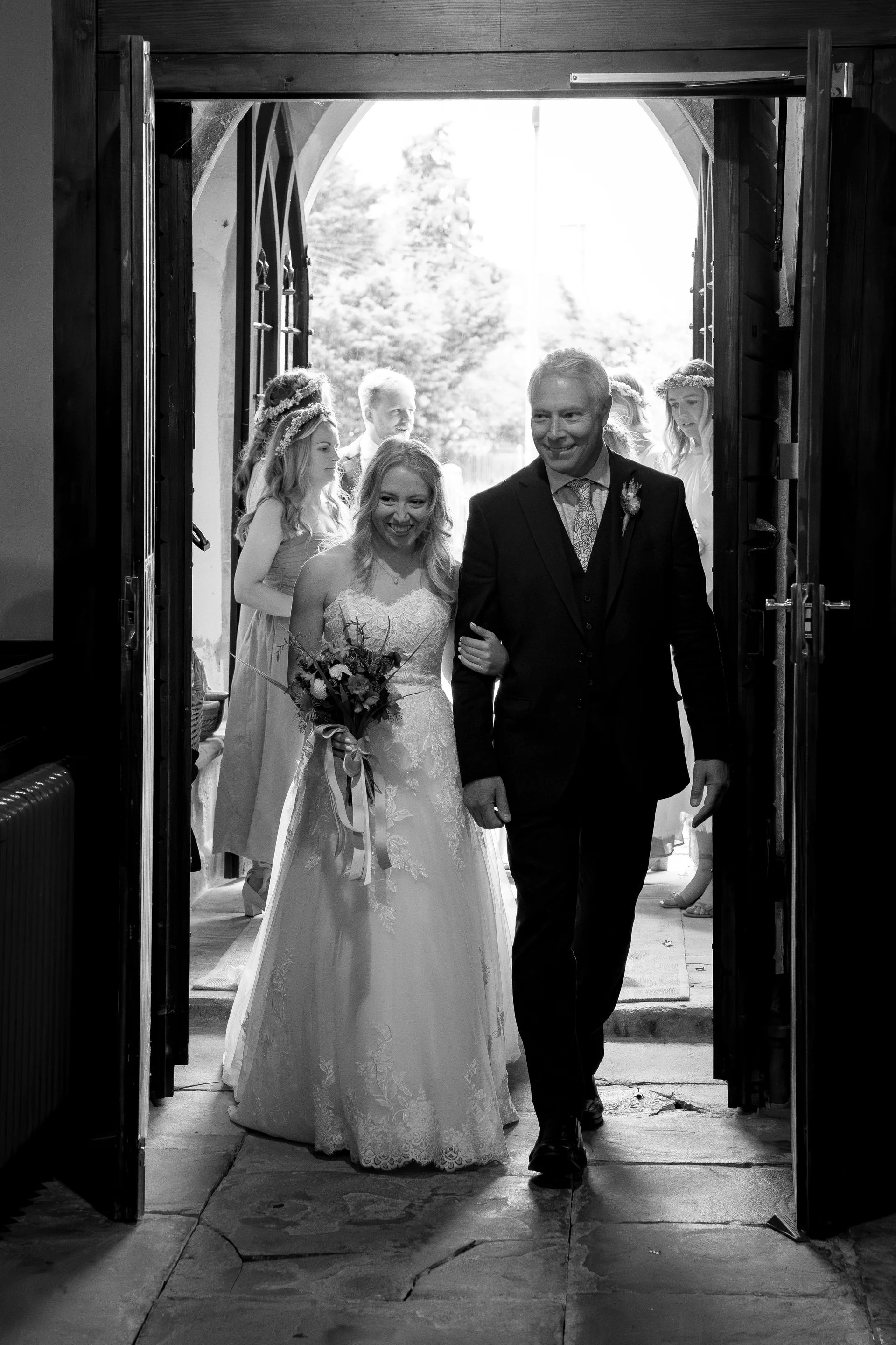 A smiling bride holding a bouquet enters a church arm in arm with her father through a large wooden doorway, accompanied by bridesmaids.