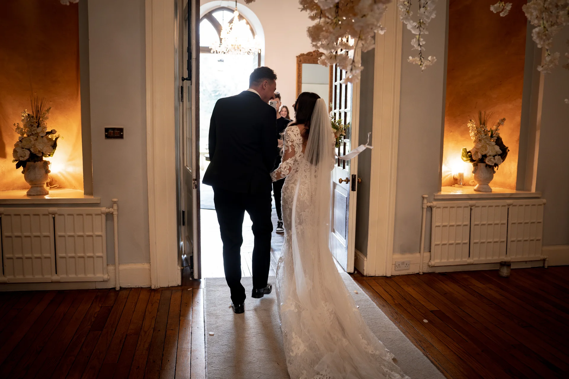 Newlyweds walk away from the camera through a bright doorway inside an elegant country house venue, with the bride wearing a long-sleeved lace gown and veil.