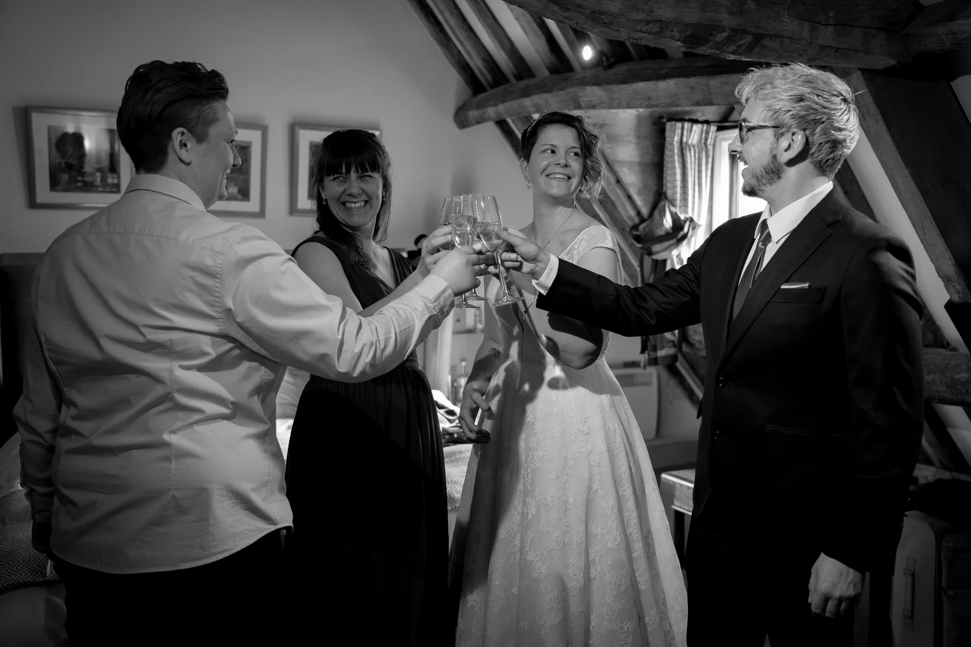 A wedding party and guests share a joyful champagne toast inside a rustic barn with exposed wooden beams, captured in a candid black and white photograph.