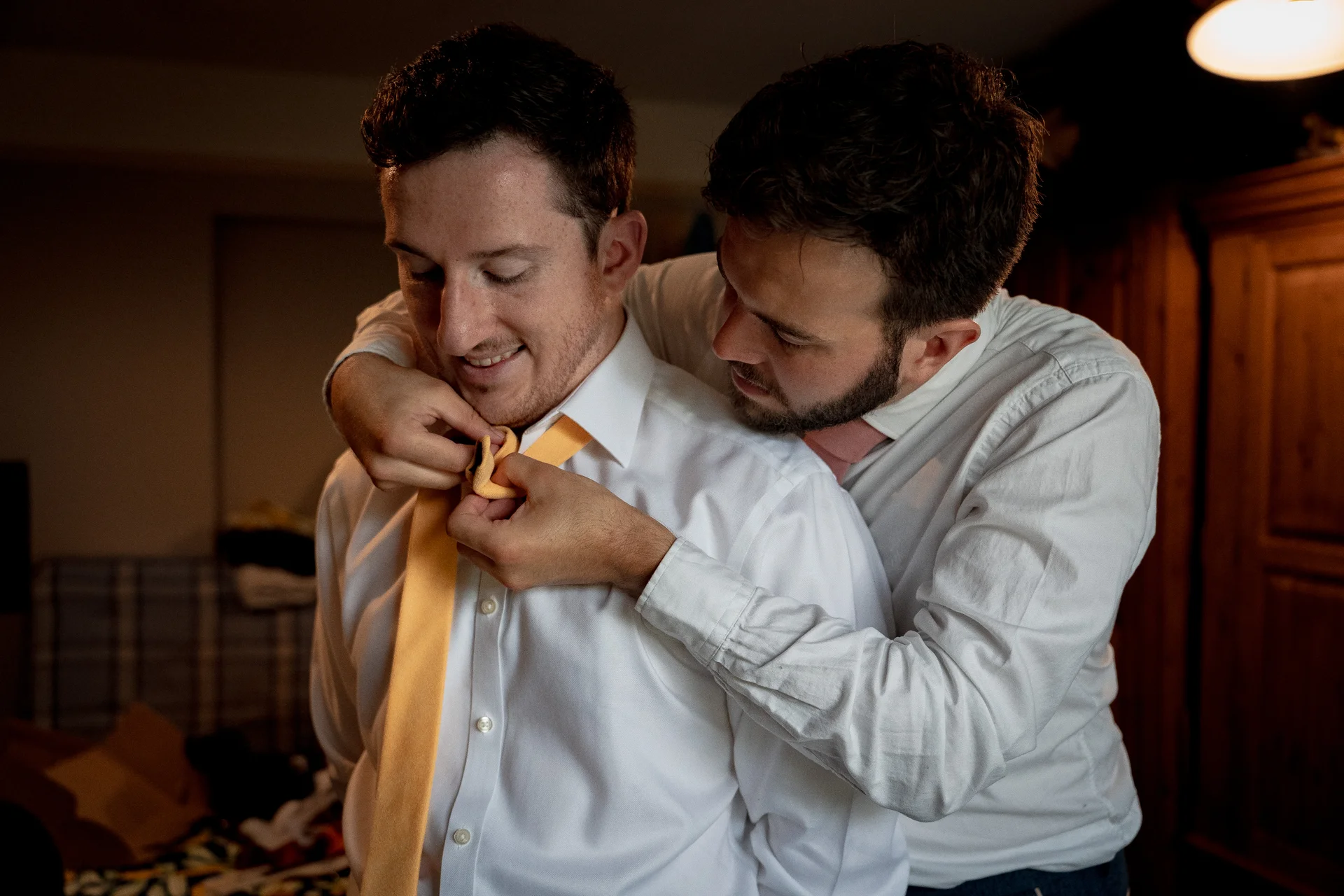 One groom gently helps tie a bright yellow tie for his partner during morning preparations in a warm, intimately lit room.