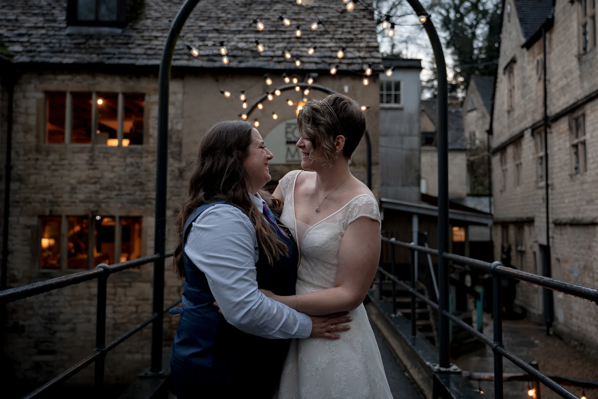 Two brides embrace lovingly on a stone bridge framed by glowing fairy lights outside a historic building at dusk.