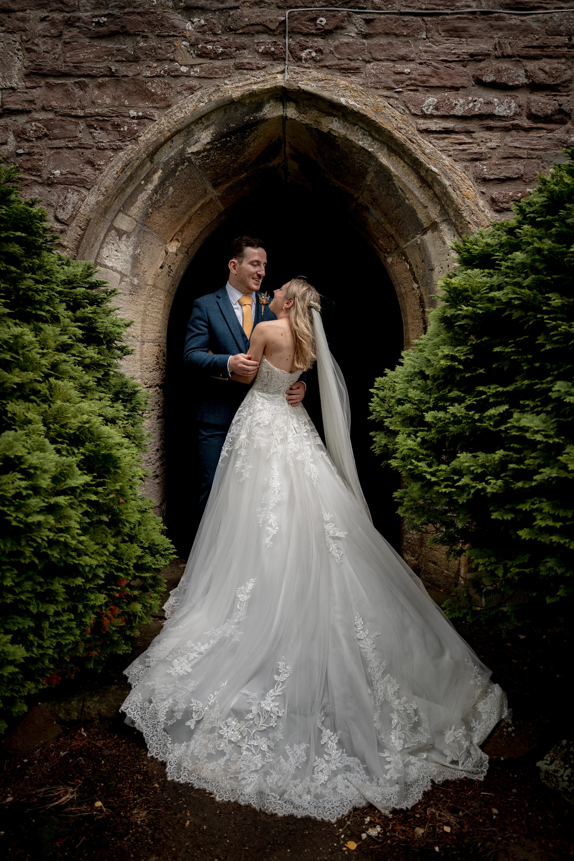A bride and groom share a tender moment standing inside a historic stone church archway flanked by lush green bushes.