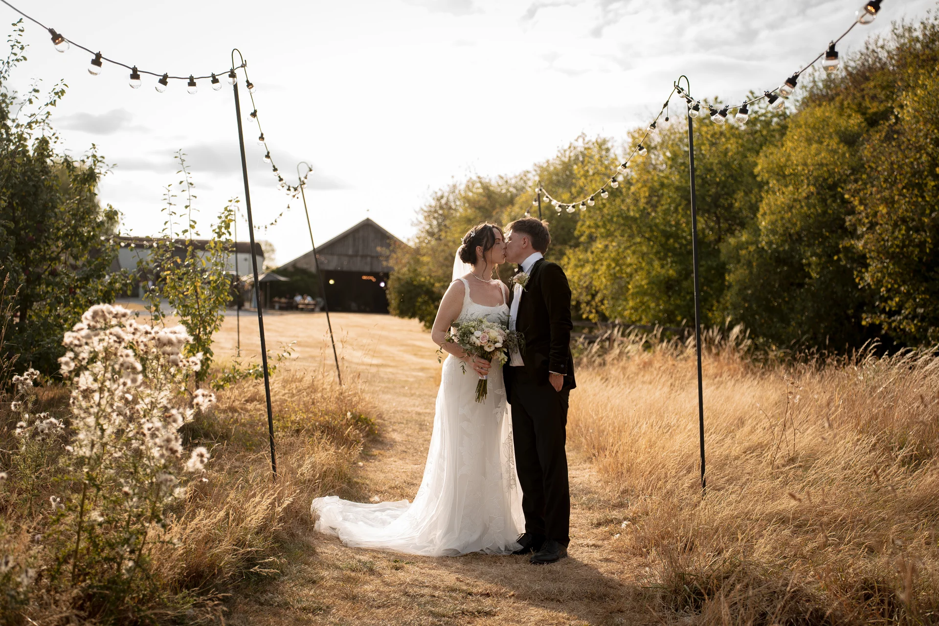 Newlyweds share a tender kiss along a rustic pathway framed by festoon lighting in a sunlit meadow with a barn in the background.