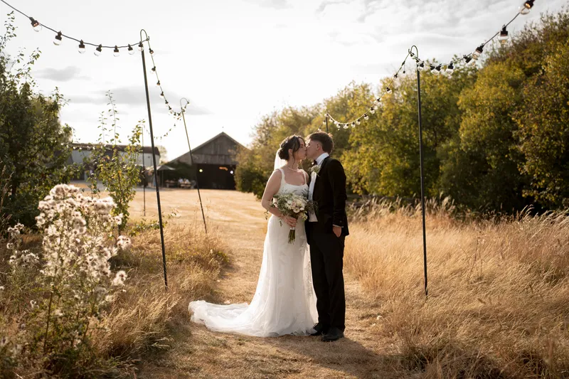 Newlyweds share a tender kiss along a rustic pathway framed by festoon lighting in a sunlit meadow with a barn in the background.