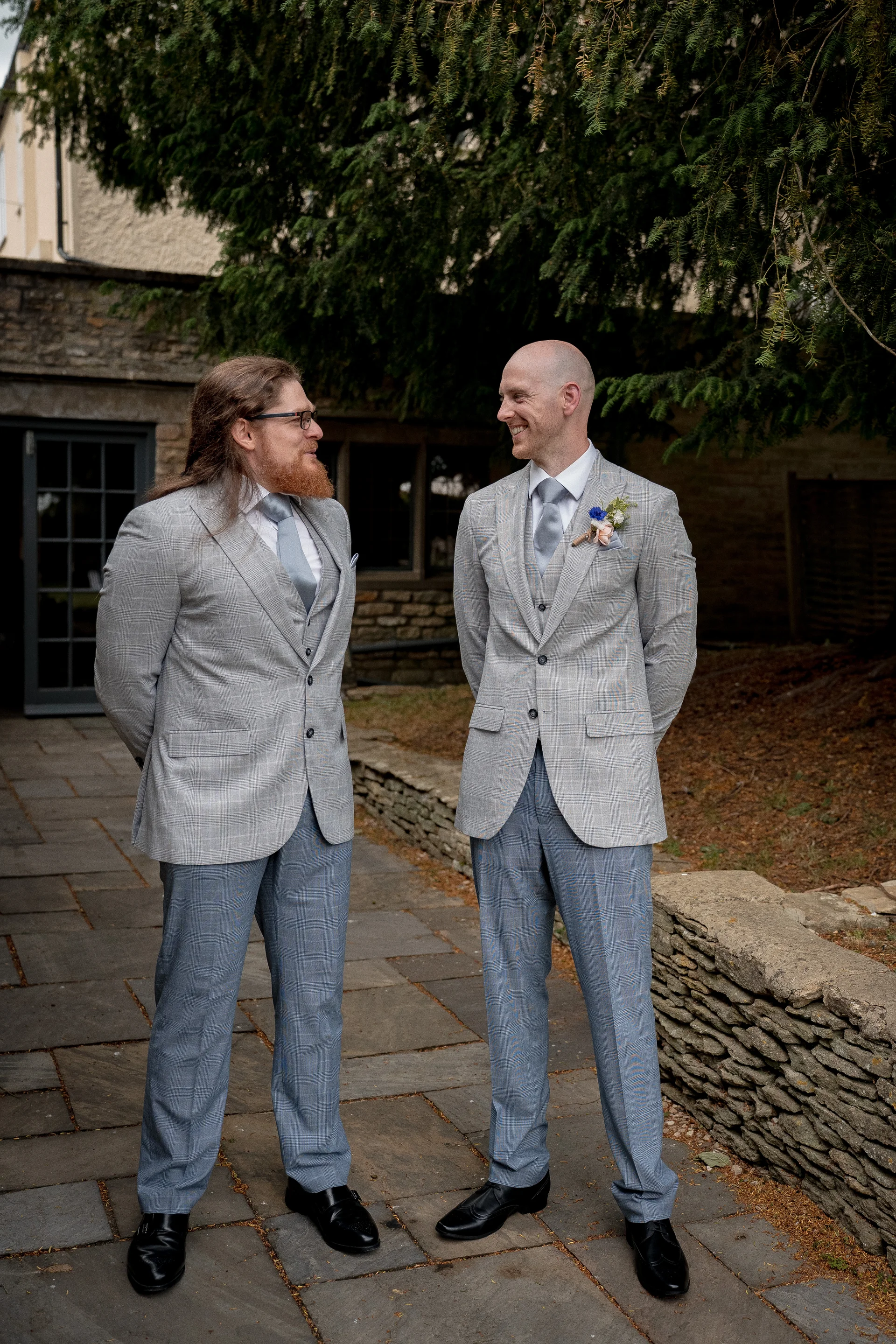 Two grooms in matching grey checked suits and ties stand smiling at each other in a stone courtyard.