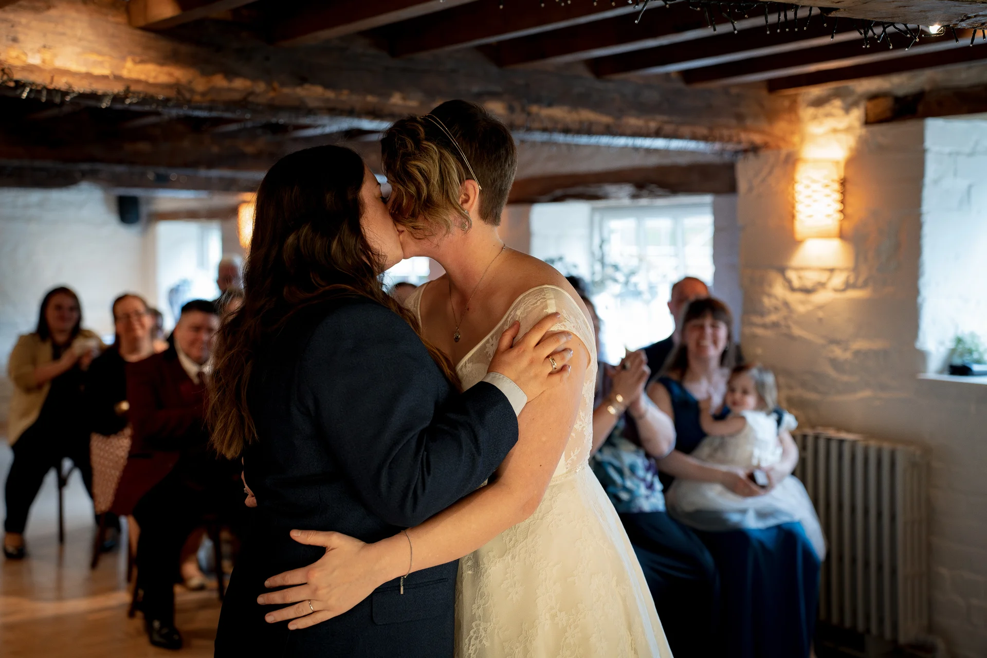 Two brides share a tender first kiss during their rustic barn wedding ceremony while guests watch and applaud in the background.