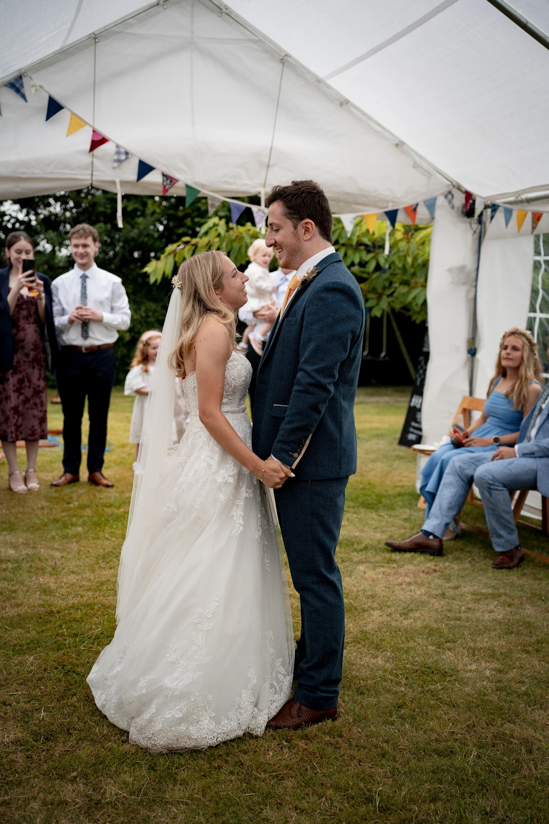 A bride in a lace a-line gown and groom in a blue tweed suit hold hands during an outdoor marquee ceremony with bunting overhead.
