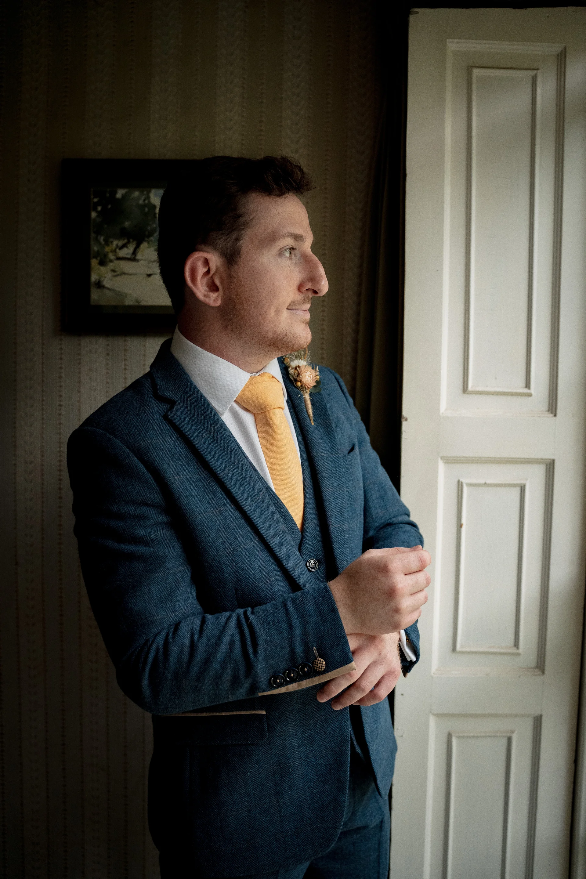 A groom in a blue patterned three-piece suit and a yellow tie adjusts his cufflinks while standing near a panelled doorway in natural window light.