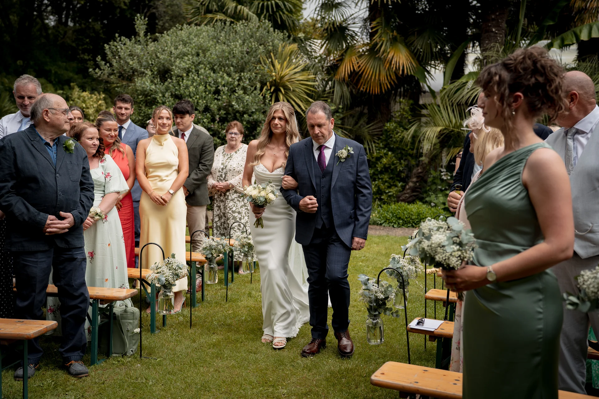 A bride walks down the outdoor aisle linked arm-in-arm with her father, holding a white bouquet while guests seated on wooden benches look on.
