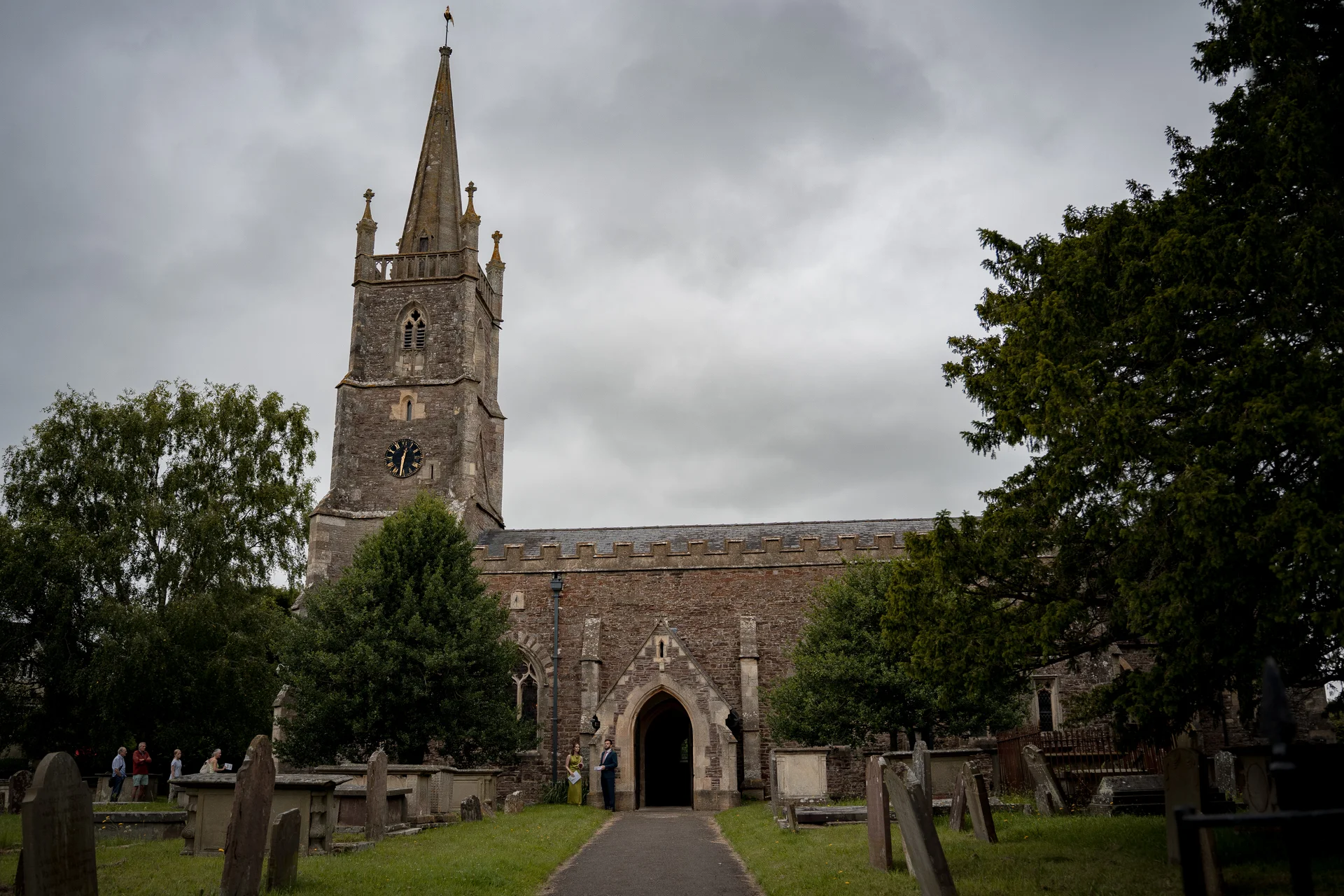 A tiny couple stands outside the stone archway of an ancient historic church with a tall steeple under a moody overcast sky.
