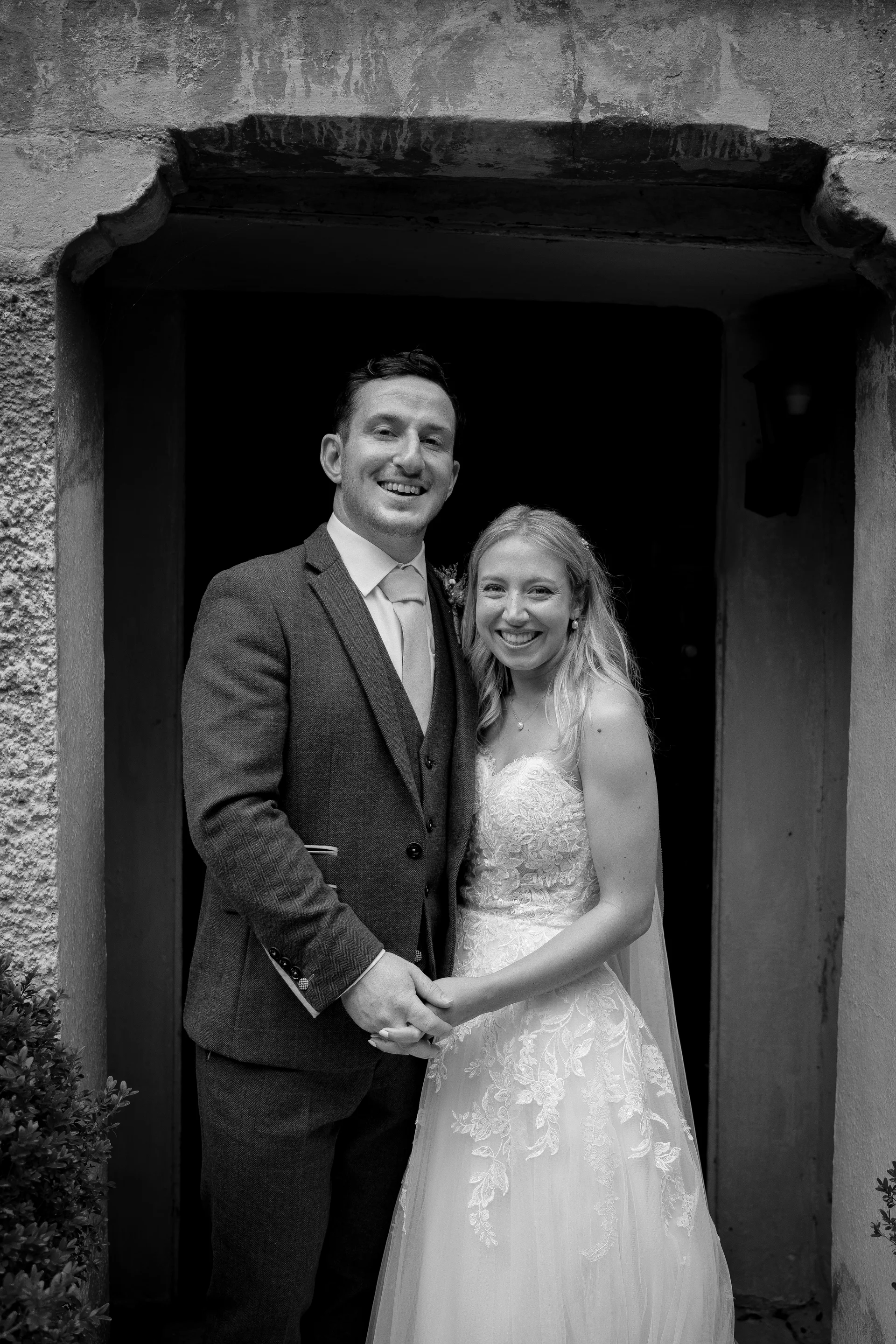 A happy bride and groom stand smiling together, framed by a traditional stone doorway in this classic black and white wedding portrait.