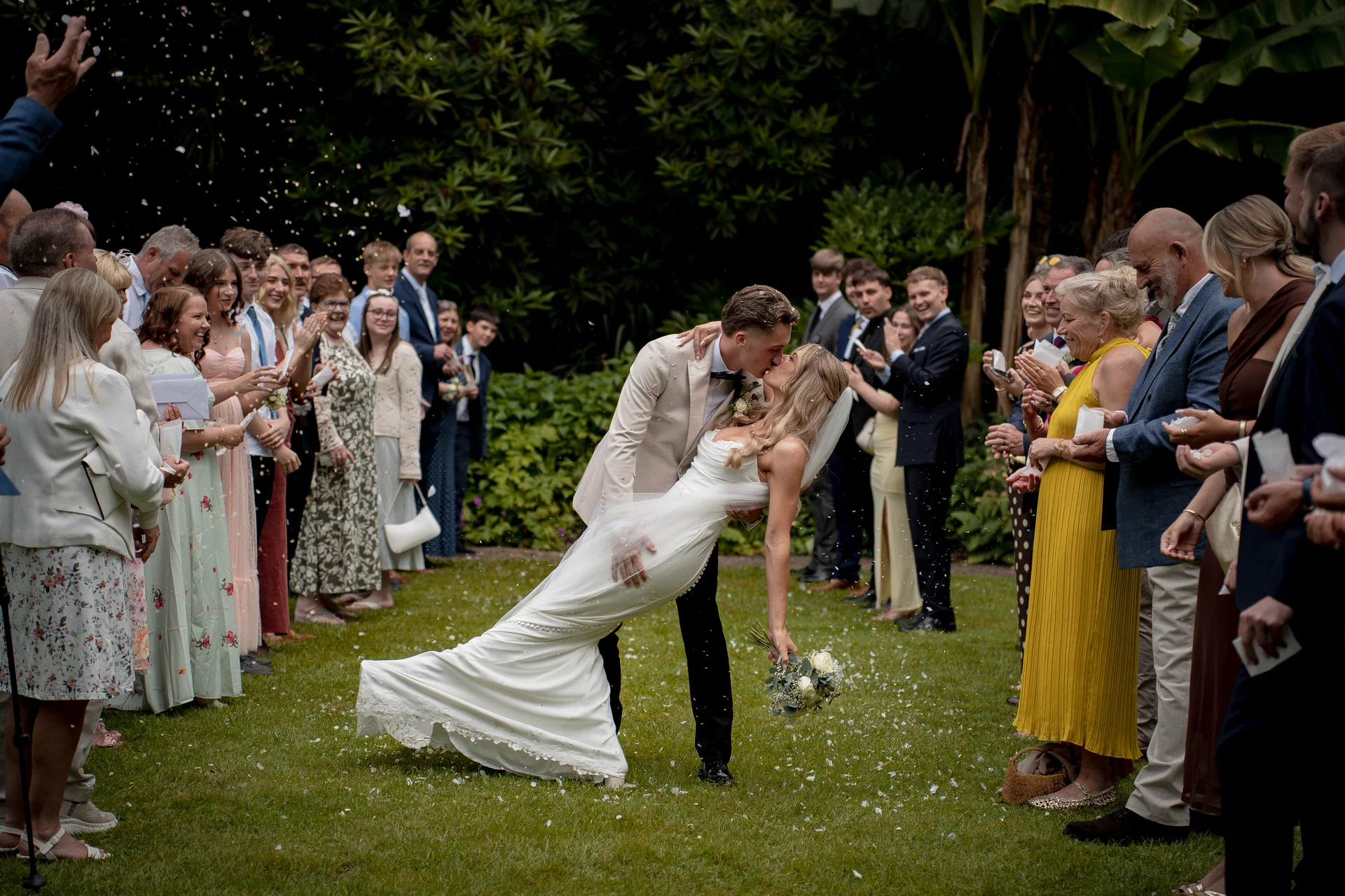 Newlyweds share a dramatic dip kiss as cheering guests shower them with confetti on a lush green lawn.