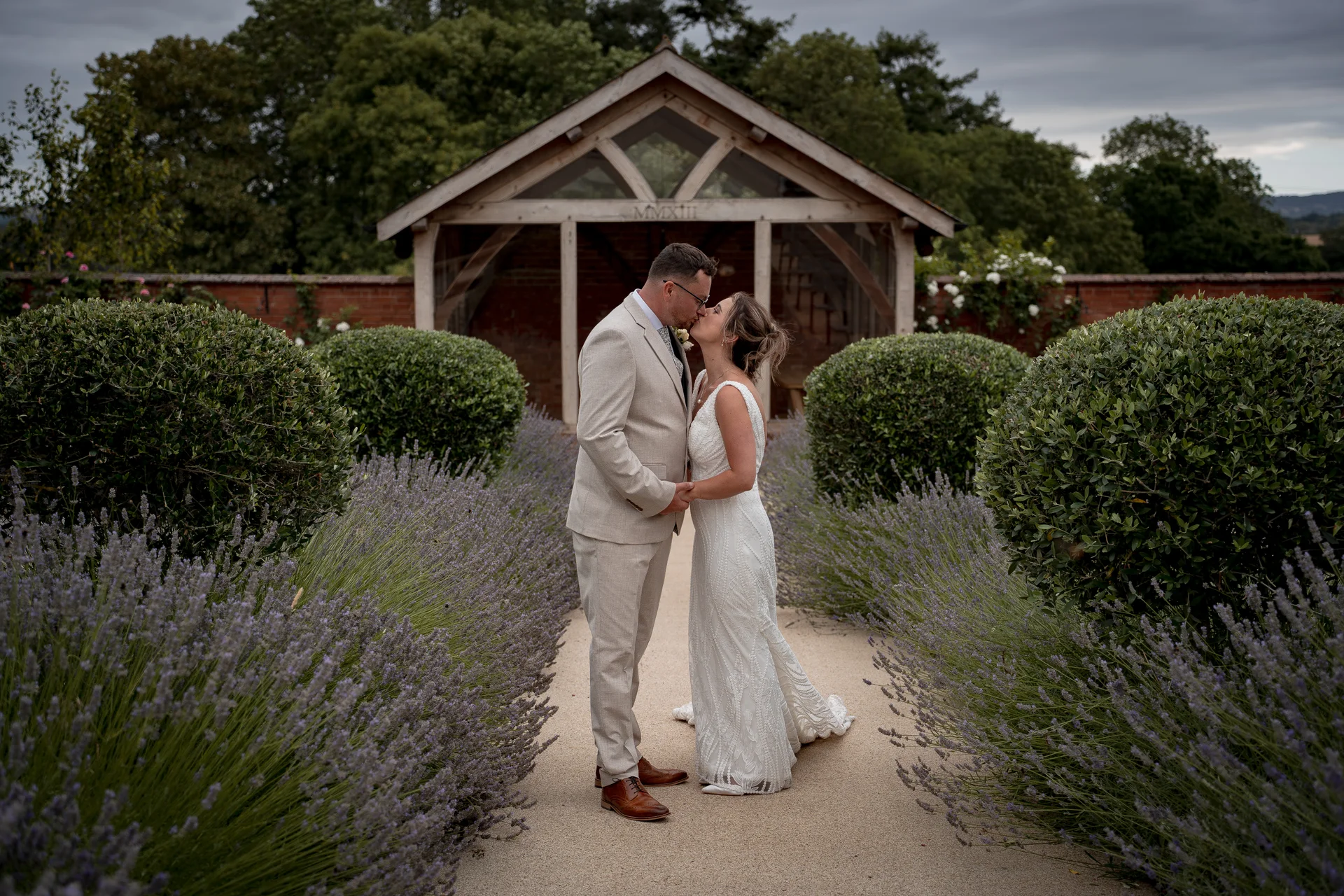 A newlywed couple shares a tender kiss along a gravel pathway lined with blossoming lavender beds and manicured topiary bushes in an English country garden.