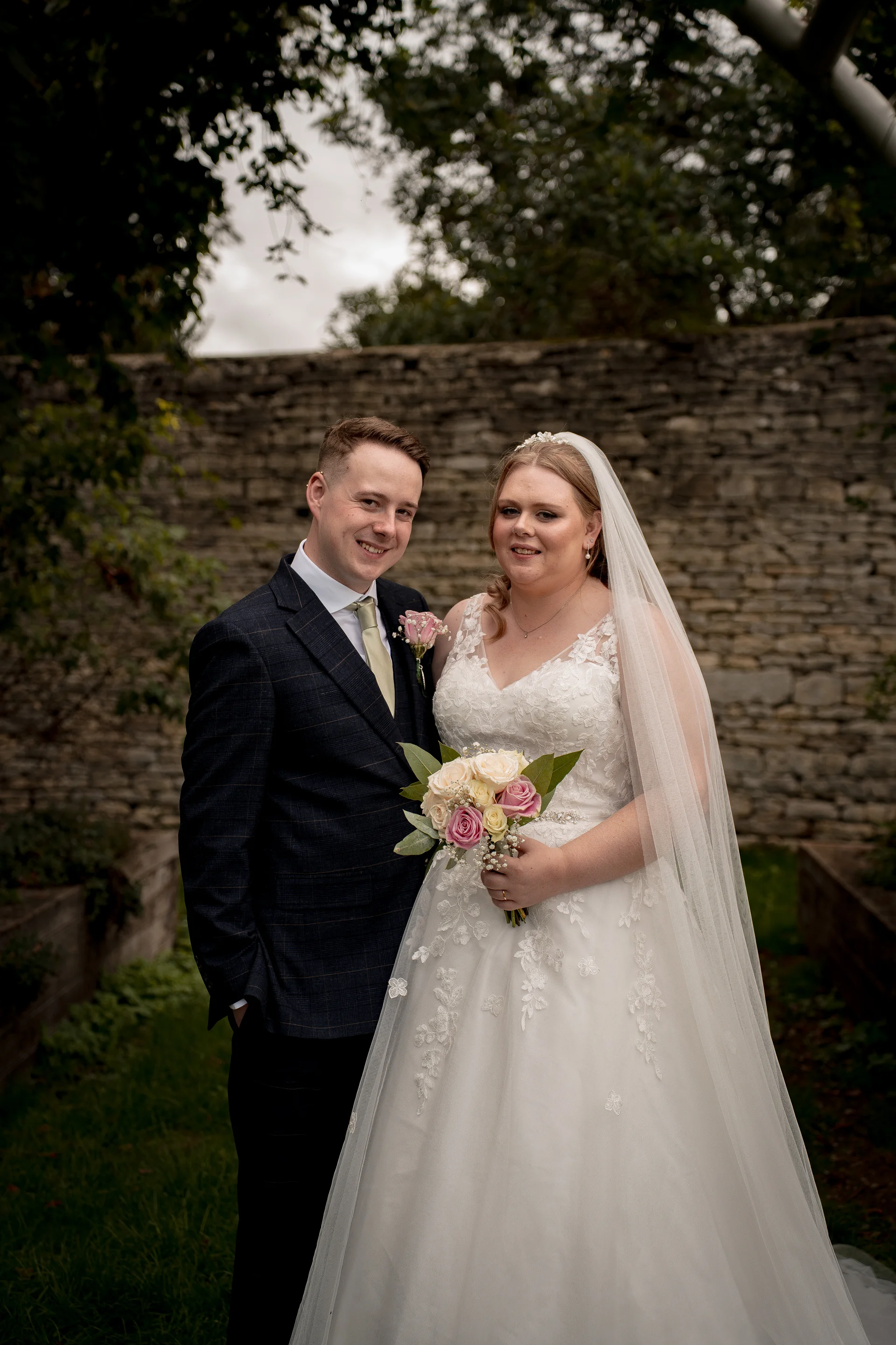 A smiling bride and groom pose together outdoors in front of a rustic stone wall, with the bride holding a mixed rose bouquet and wearing a lace a-line gown and veil.