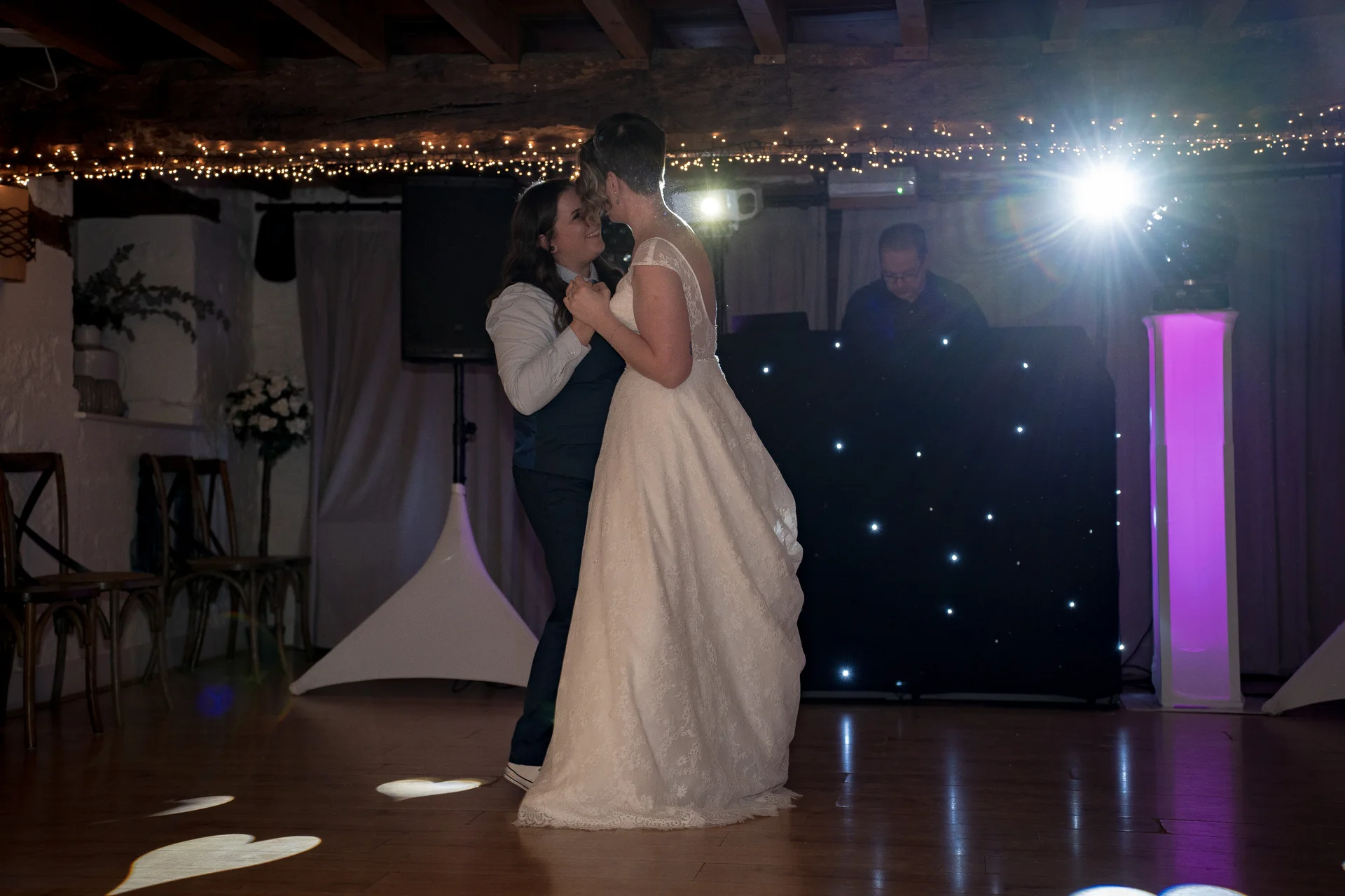 Two brides share their first dance together inside a rustic barn venue illuminated by twinkling fairy lights and stage lighting.