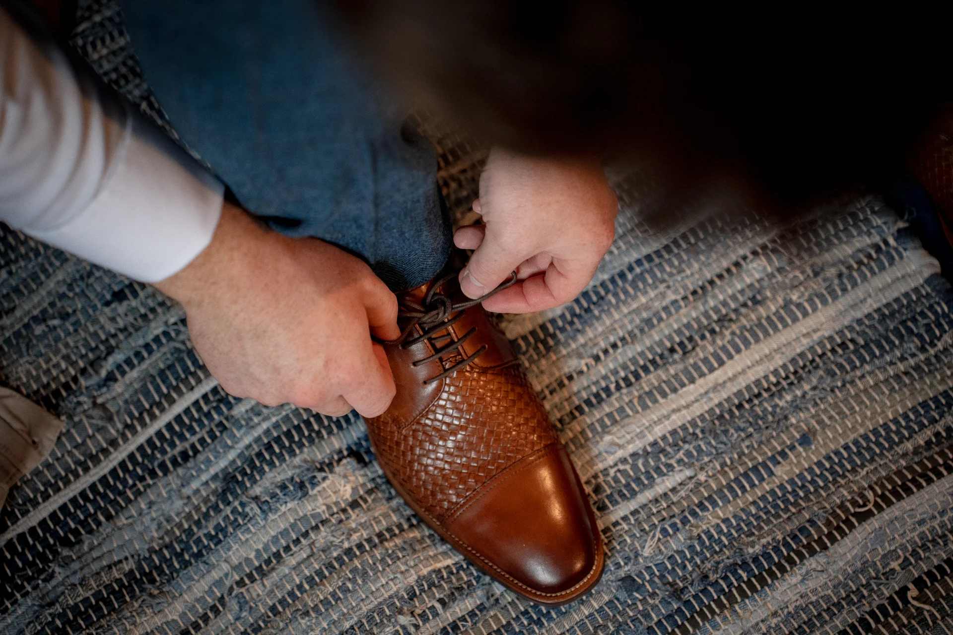 A groom carefully ties the laces of his brown leather woven brogues while getting ready on a rustic blue and white rug.