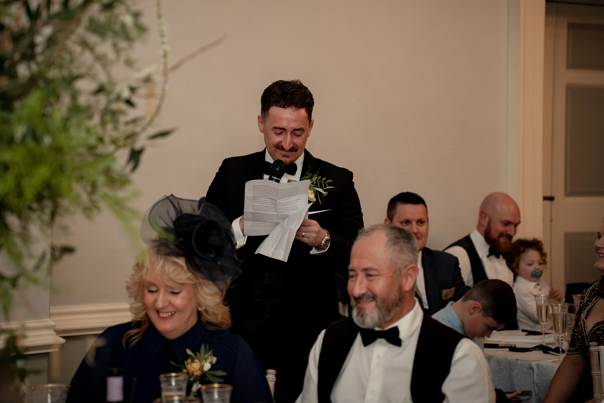 A groom in a black tuxedo delivers a speech while reading from paper notes during the wedding reception, with smiling guests seated around tables.