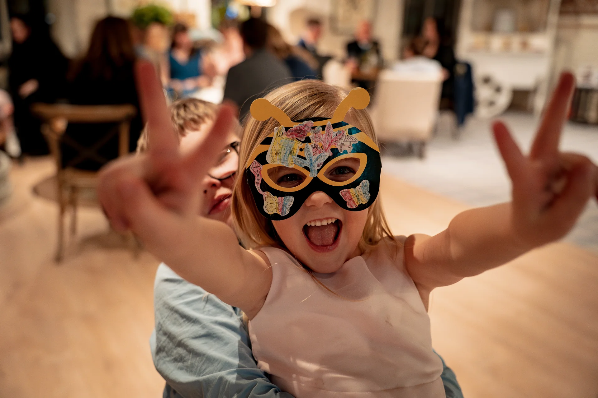 A young flower girl wearing a decorative paper mask excitedly throws her hands up in a peace sign while being held by a page boy at a wedding reception.
