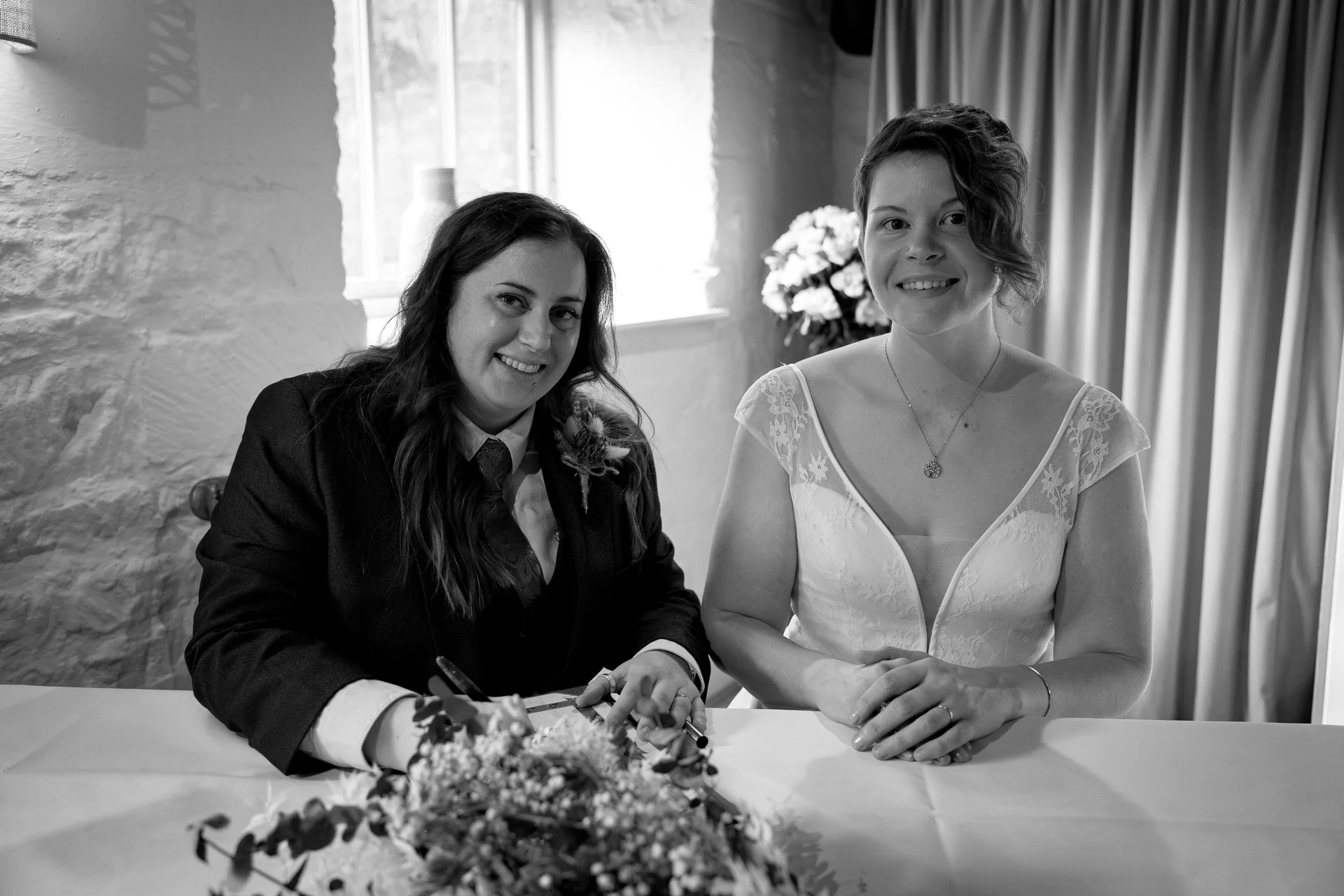 Two brides sit smiling side by side at a table while signing the register during their wedding ceremony.