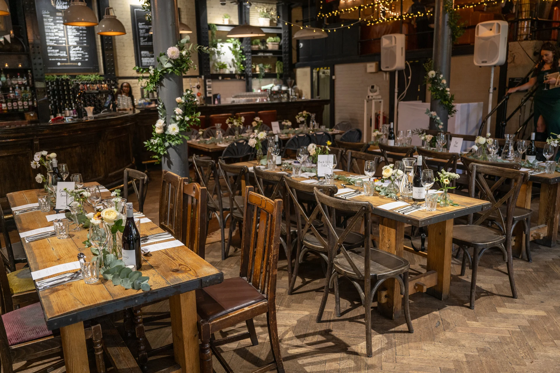 An industrial warehouse wedding venue interior set up with long wooden banquet tables featuring cross-back chairs, greenery runners, and delicate bud vase arrangements.