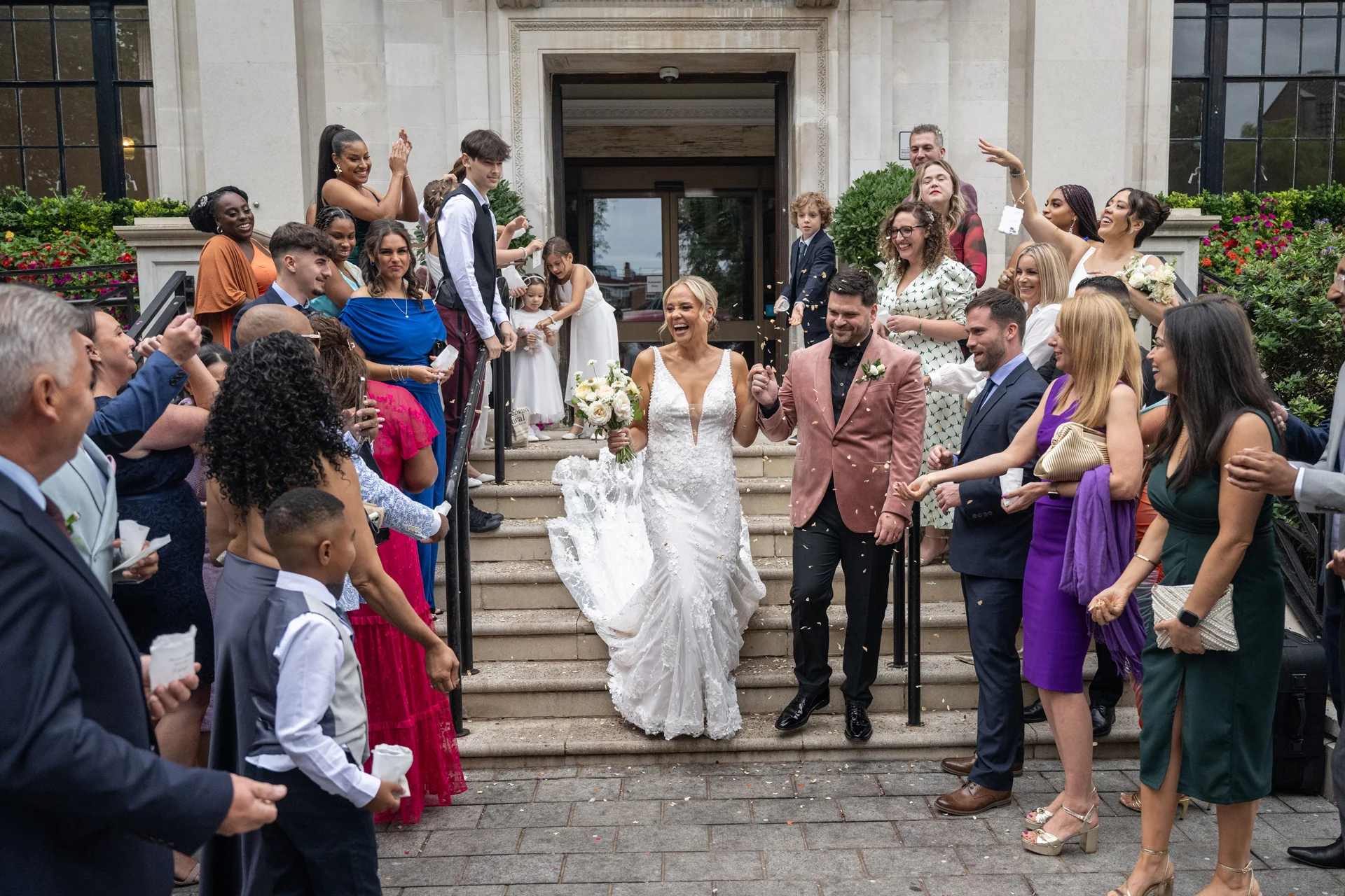 A joyful newlywed couple exits the venue hand in hand down stone steps as cheering wedding guests shower them with confetti.