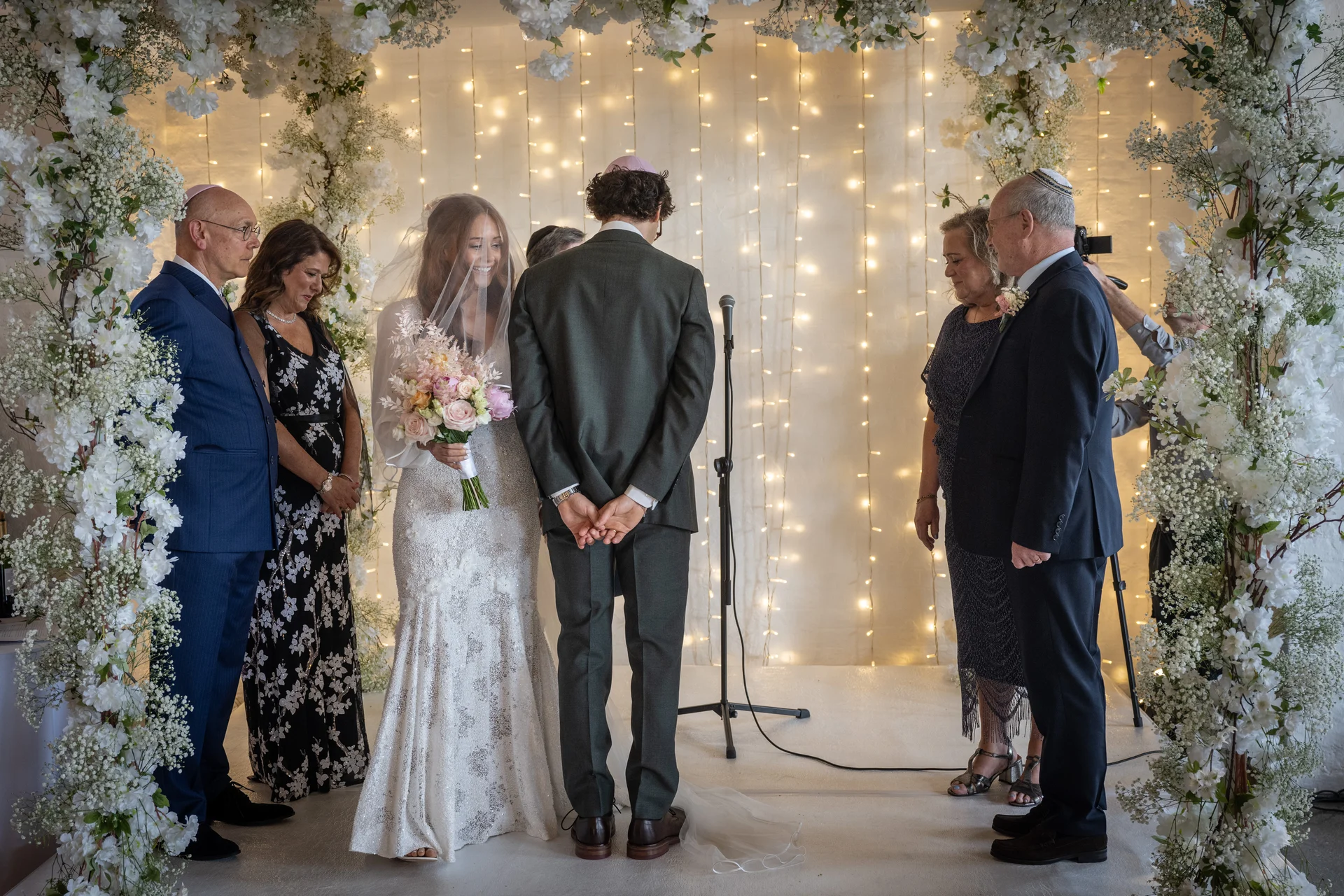 A bride in a lace dress with a blusher veil and a groom in a green suit stand beneath a floral archway during a candlelit ceremony surrounded by family.