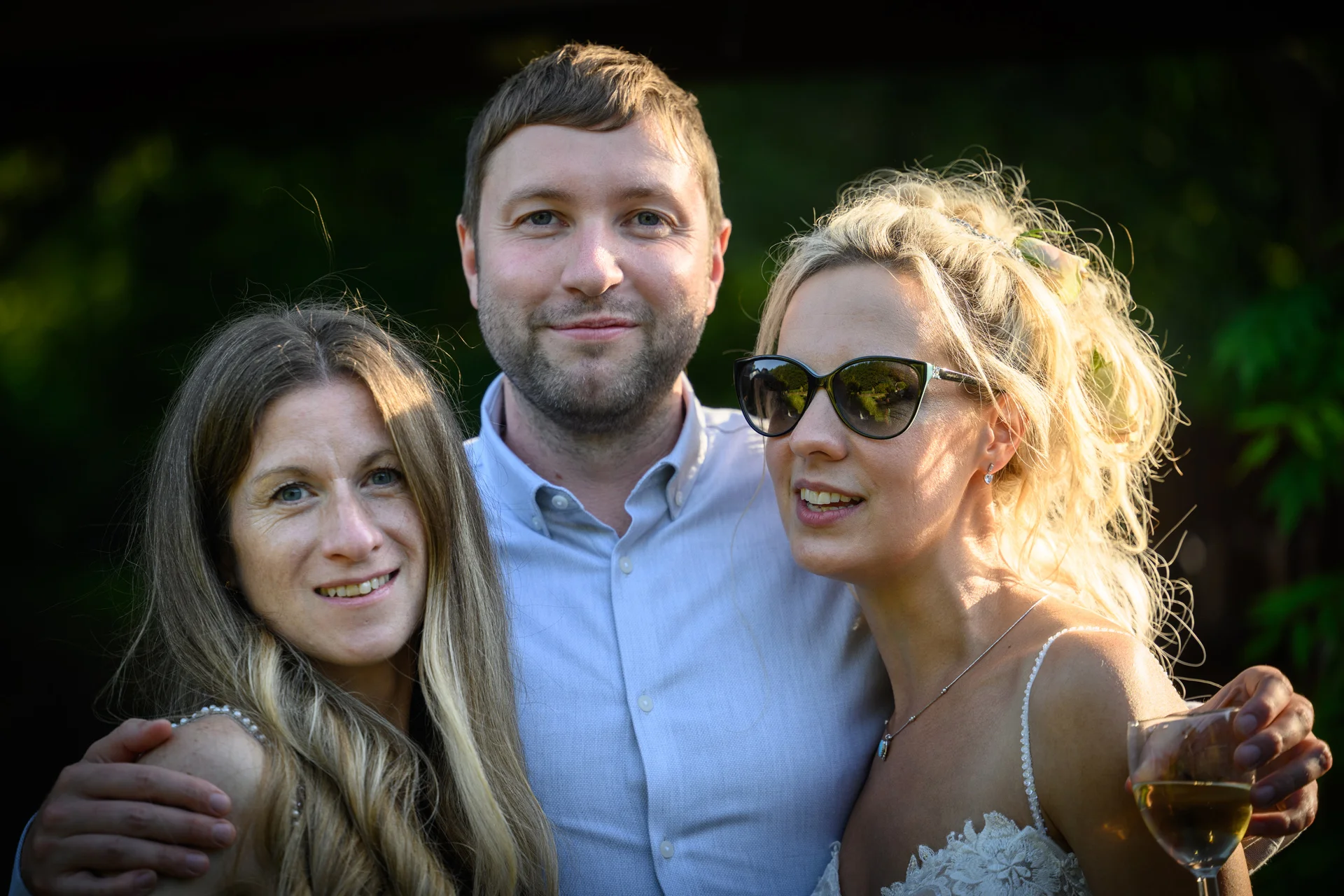 A smiling couple and a female guest pose closely together outdoors during the golden hour, with the bride wearing sunglasses and holding a glass of wine.
