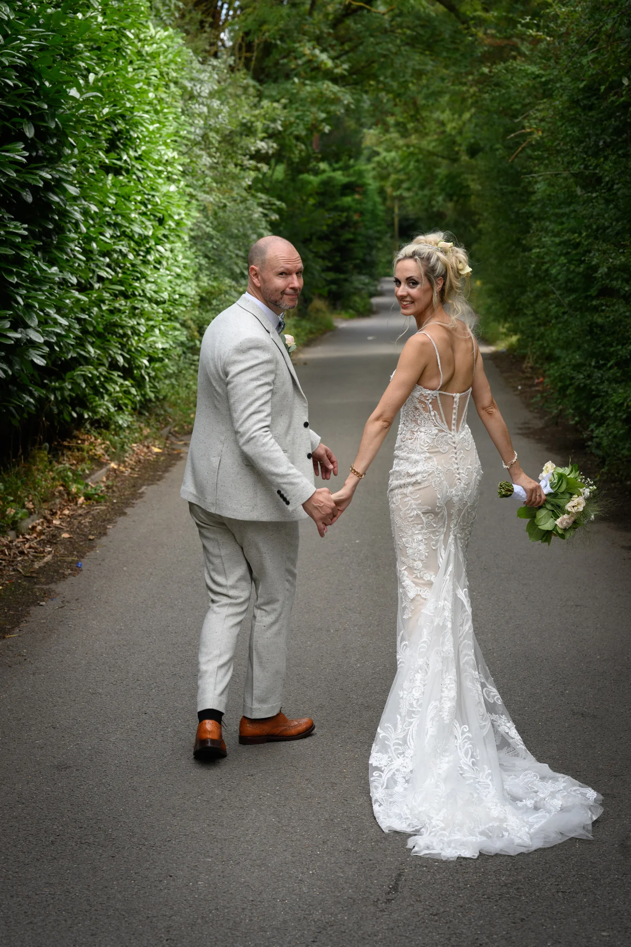 A newlywed couple walking hand in hand down a tree-lined lane look back over their shoulders at the camera, with the bride holding a delicate bouquet.