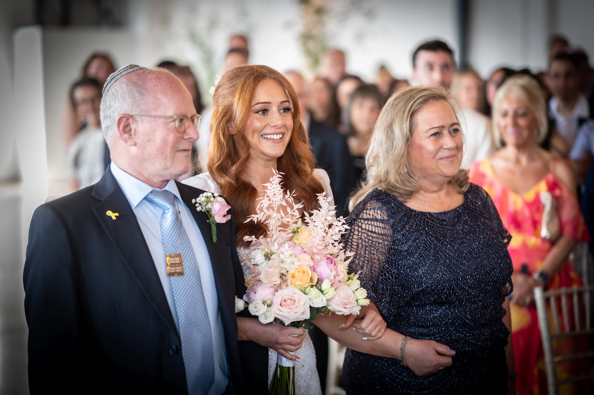 A smiling red-haired bride walks down the aisle arm in arm with her parents during the wedding ceremony.